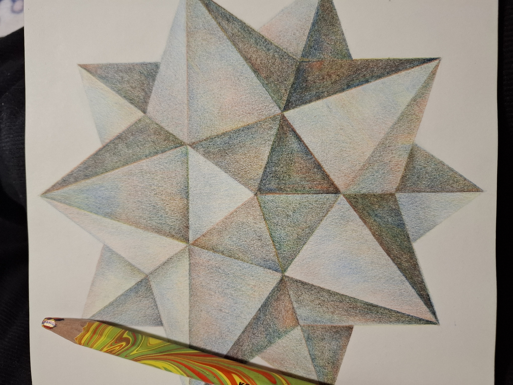 A close-up shot captures a colored pencil drawing of a geometric star shape on white paper. The drawing is rendered with subtle shading, creating a sense of three-dimensionality. A multi-colored pencil, featuring swirls of yellow, red, and green, rests on the left side of the paper, partially over the drawing. The background appears to be a dark fabric. There is no visible indication of the city of Tyumen, Russia, or any specific urban environment in this image.