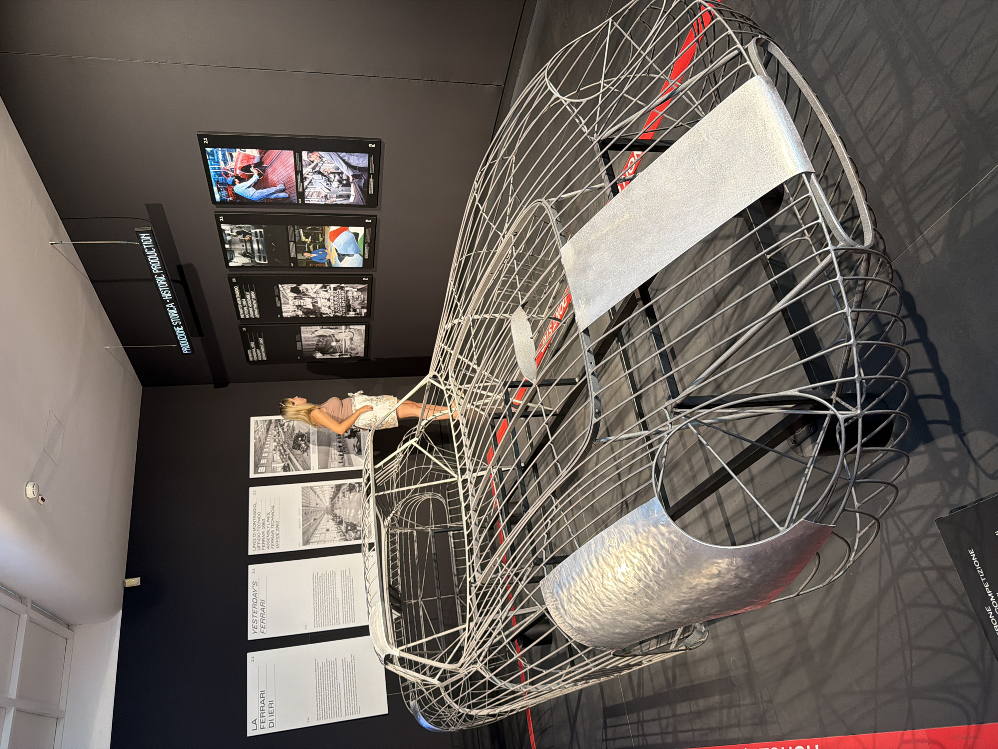 The image depicts a wireframe sculpture of a classic Ferrari car, likely a representation of its chassis. The sculpture is situated indoors, possibly in a museum or exhibition space, with a dark gray wall as the backdrop. To the left of the sculpture, there are three large informational posters with text and images. A woman, wearing a light-colored top and a floral skirt, is standing near the posters, looking at the car sculpture. Above the posters and to the right, a neon sign displays "PRODUZIONE STORICA - HISTORIC PRODUCTION" in white lettering. Further to the right, there are four framed displays containing historical photographs or images related to Ferrari production. The floor is dark and reflective, showing shadows cast by the sculpture. The overall environment suggests a formal exhibition dedicated to the history of Ferrari. Given the context of Ferrari, the location is likely Maranello, Italy, the home of Ferrari.