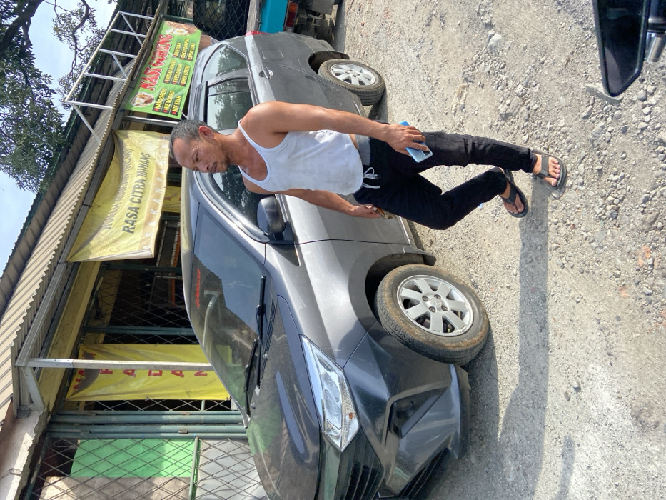 The scene is a street in Jatiluhur, Indonesia (latitude: -6.577, longitude: 107.436). A man in black pants and a white tank top stands next to a grey Toyota Avanza, which is parked on a gravel surface in front of a building. The building has a yellow sign that reads “RASA CITRA MINANG.” There is a second sign with Indonesian text on it. The man is holding a blue phone in his right hand. The Avanza’s driver’s side door is open. The man is looking to the left, towards the building. The sky is visible in the upper right corner of the photo. The ground is a mix of gravel and dirt. A small piece of the rearview mirror of another vehicle is visible in the lower right corner. The man is wearing sandals. The Avanza’s front tire is facing directly toward the camera. The man is standing a short distance from the Avanza.