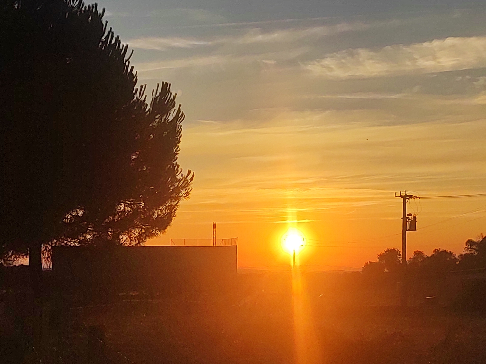 The image captures a sunset in Sobradillo, Spain, characterized by a vibrant sky filled with hues of orange, yellow, and pale blue. The sun is positioned low on the horizon, its bright light creating a lens flare effect that radiates downwards. In the foreground, a silhouetted coniferous tree stands on the left, its dark foliage contrasting with the bright sky. To the right of the tree, a low-rise building with a railing on its roof is visible, also silhouetted against the sunset. Further to the right, a utility pole with electrical equipment stands prominently, with a line of silhouetted trees forming the background. The overall scene depicts a rural or semi-rural landscape at dusk.