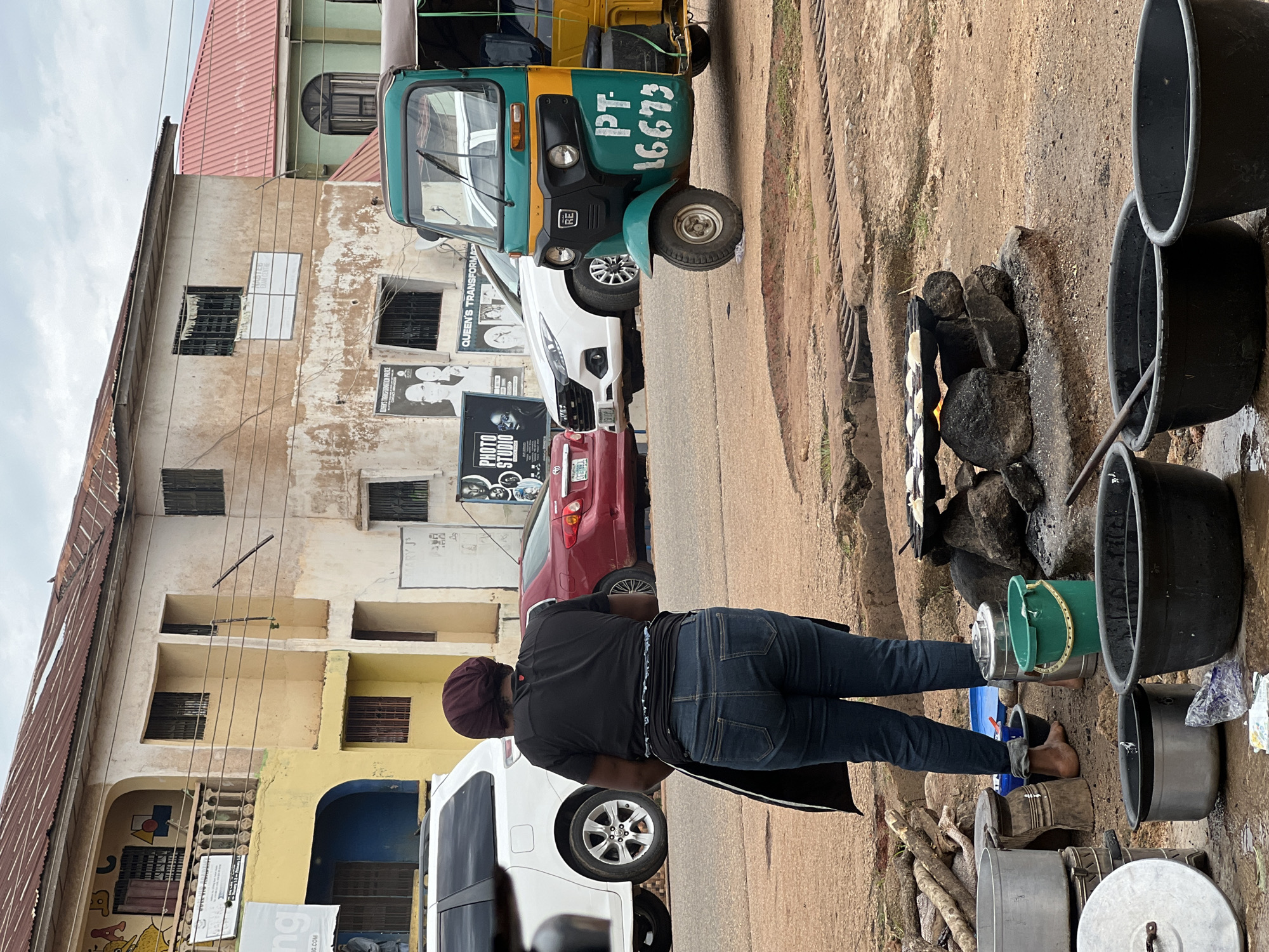 In the city of Jos, Nigeria, on a cloudy day, a woman stands in front of her food stall, preparing to cook. She wears a dark t-shirt, blue jeans, and a maroon headscarf, and her back is to the camera. In front of her, a collection of cooking utensils, including pots, pans, and buckets, are arranged on the ground. A makeshift stove, made of rocks and burning wood, is actively cooking small circular items on a flat pan. Behind her, a bright yellow and green auto-rickshaw is parked on the street, along with a white SUV and a red sedan. The buildings in the background appear weathered and aged, with some signs of disrepair. The scene suggests a typical street-side food vendor in Jos, Nigeria.