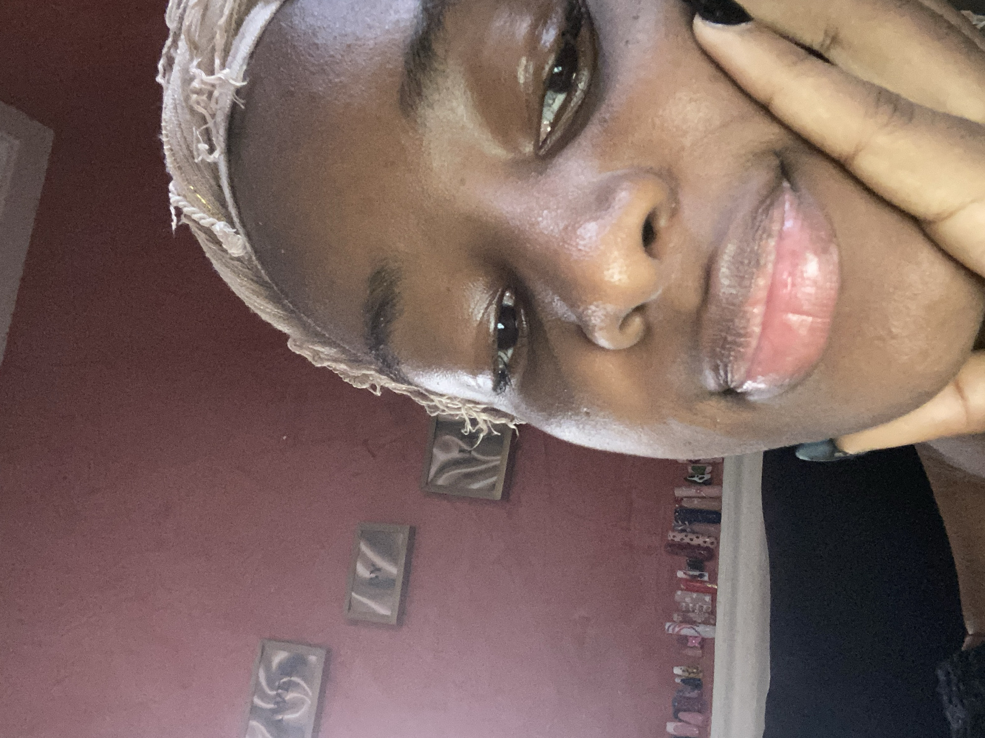 The image is a close-up, head-and-shoulders shot of a young Black person, taken from a low angle. The subject's head is tilted, and their right hand is resting on their cheek and forehead. They are wearing a light-colored headscarf with frayed edges.
The subject's skin appears dewy and reflective, suggesting a natural glow or the application of a hydrating product. Their eyes are partially visible, with one eye looking towards the camera and the other slightly averted. Their lips are full and have a glossy finish.
The background is out of focus but appears to be an indoor setting. The wall behind the subject is a muted pinkish-red color. Several small, framed pictures or artwork are mounted on the wall, arranged vertically. To the right, a white curtain or fabric is visible, and further to the right, a stack of colorful items, possibly books or decorative objects, are arranged vertically. The lighting suggests it is daytime, with natural light illuminating the subject's face.
The overall mood is intimate and self-reflective, with the subject's pose and expression conveying a sense of calm or contemplation. There is no visible text in the image. The location context provided is Jos, Nigeria.