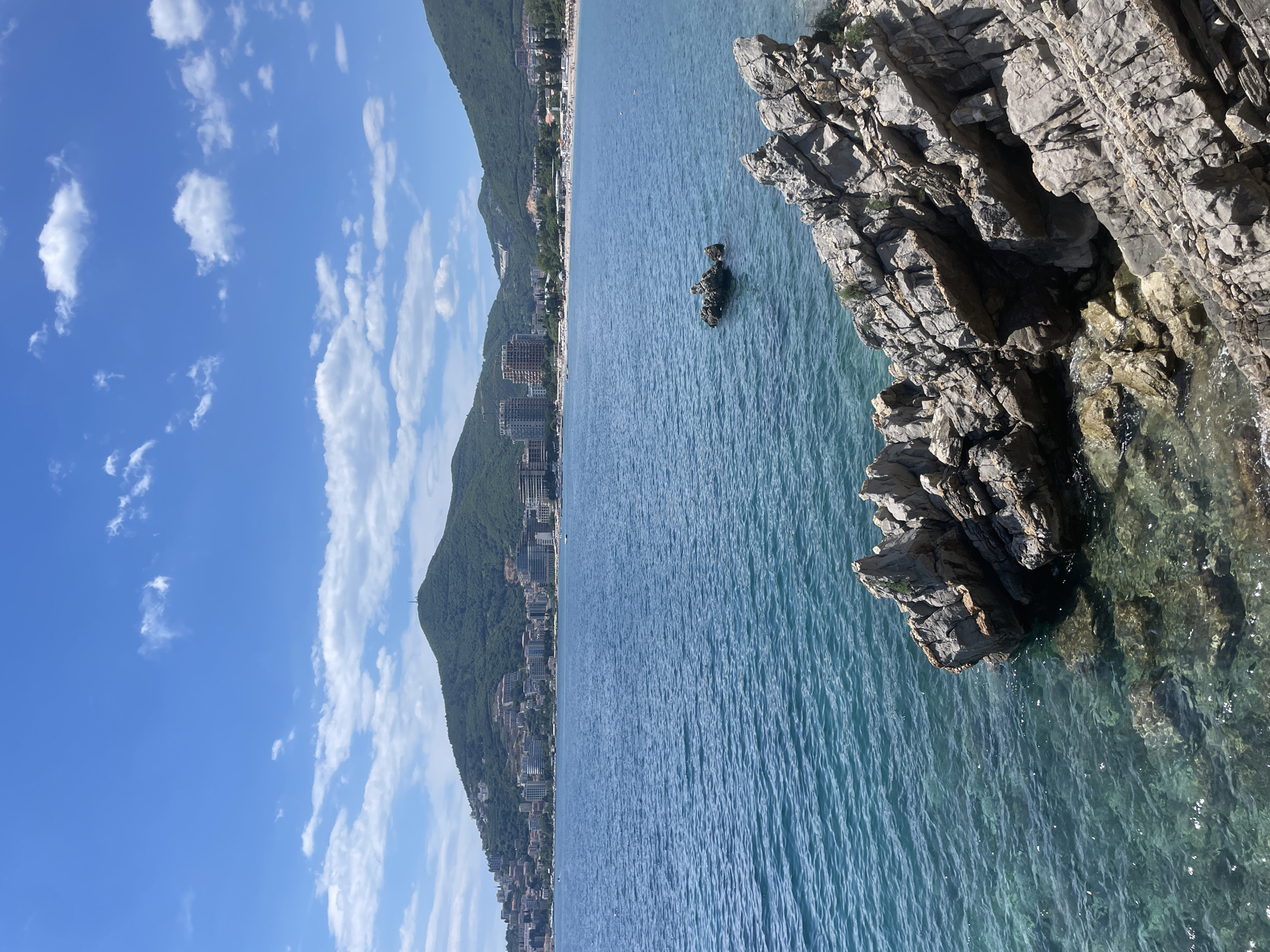 This image depicts the coastline of Budva, Montenegro. A rocky cliff dominates the right side, descending into the clear, turquoise water of the Adriatic Sea. The water is calm with visible wave patterns. In the distance, the city of Budva is visible, built along the base of green, mountainous terrain. A blue sky with scattered clouds is overhead.