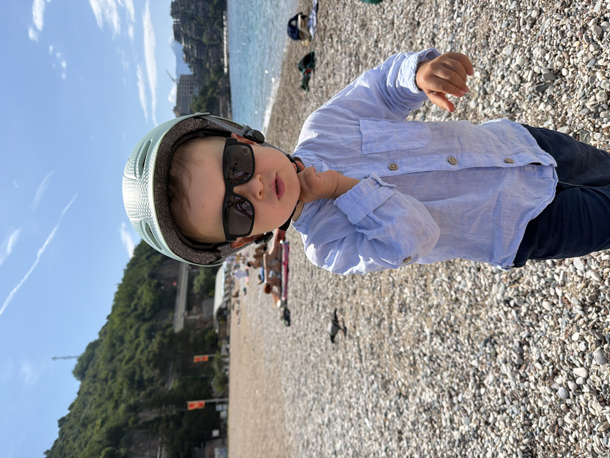 A young child wearing a light blue helmet and sunglasses stands on a pebble beach. The child wears a light blue button-down shirt and dark pants. Behind the child, the beach is visible, along with other people. In the background is the Adriatic Sea and a hillside covered in trees. The scene depicts Boreti, Montenegro.