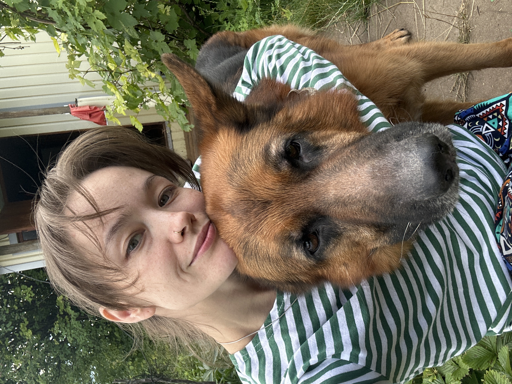 A young woman with light brown hair and a nose ring takes a selfie with a German Shepherd. She is wearing a green and white striped t-shirt and a necklace. The dog is wearing a green and white striped bandana around its neck and is nuzzling into the woman's chest. The background appears to be a yard with trees and a light-colored building.