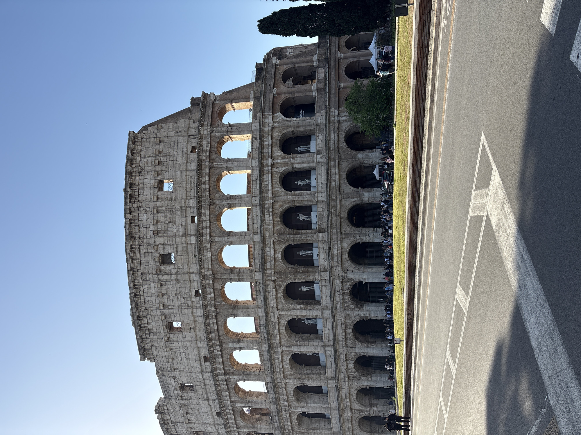 The image displays a rotated, portrait-oriented view of the Colosseum in Rome, Italy, under a clear, bright blue sky. The iconic ancient Roman amphitheater, constructed from stone, occupies the lower and left-hand portions of the frame, with its characteristic three-tiered arched facade prominently visible. Sunlight strongly illuminates the structure, casting distinct shadows.
A significant crowd of people is gathered along the base and within the lower arches of the Colosseum, indicating a busy tourist attraction. Many individuals appear to be queuing or waiting along a strip of vibrant green grass that runs vertically through the center of the image. This grassy area is bordered by a low brick wall. Small white canopy tents are also visible on the grass, suggesting visitor facilities or temporary setups.
To the right of the grassy area, an asphalt road with white painted markings, including crosswalk lines and lane dividers, fills the upper-right section of the frame. Shadows from the Colosseum or surrounding elements are cast across the road surface. Dark green trees, including a tall, slender conifer, are discernible behind the Colosseum structure in the upper left. Inside some of the upper-level arches of the Colosseum, light-colored statues or figures are visible. Faint and fragmented text, "OSSED-", is barely visible on a dark sign within one of the arches, and unreadable text appears on a small white sign near the grassy median. The bright lighting suggests a sunny day, likely morning or late afternoon, capturing a lively scene around the historic landmark.