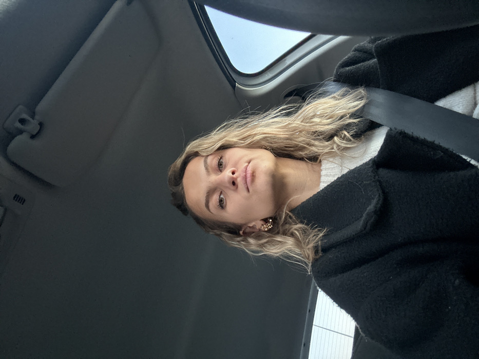 In a tilted angle portrait, a young woman is seen within a car's interior. She has light blonde, wavy hair and is wearing a black coat over a white sweater. A car seatbelt crosses her chest. The car's interior includes the sun visor and a window. The woman looks directly at the camera with a neutral expression.