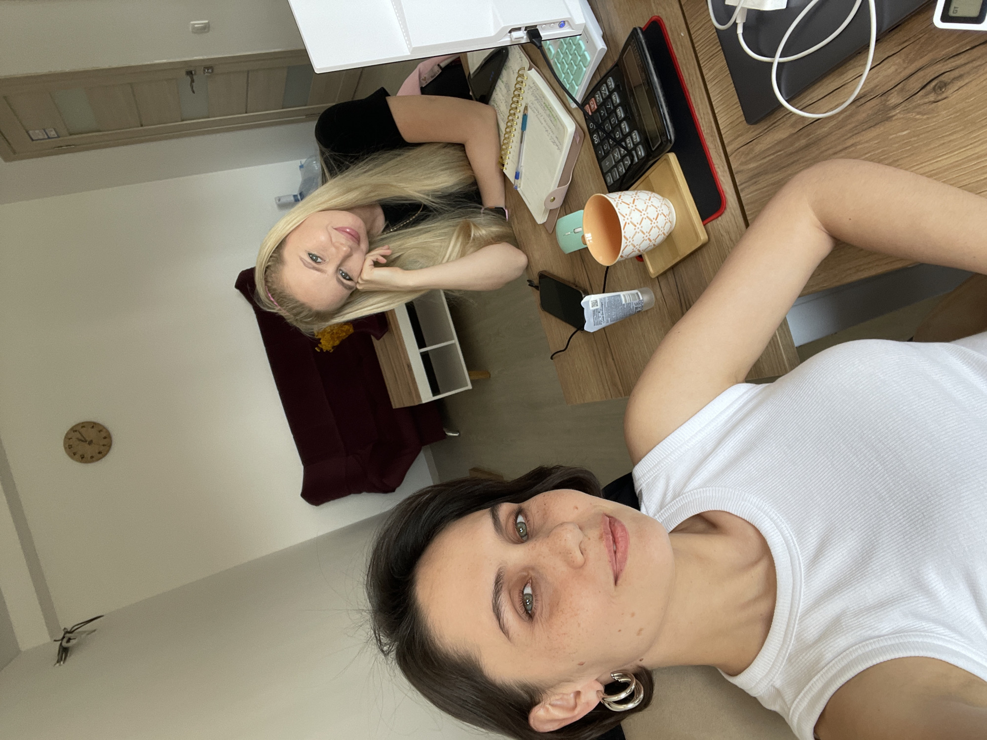 Two women are posing for a selfie in a room. The woman on the right is wearing a white top and silver hoop earrings. The woman on the left has long blonde hair and is wearing a black top. A desk separates them, which has a calculator, a mug, a notebook, and a mouse. There is a door on the top left corner of the image, and a clock on the left wall. There is a small figurine on the wall underneath the clock. The location in the city of Izhevsk, Russia, cannot be determined from the image.
