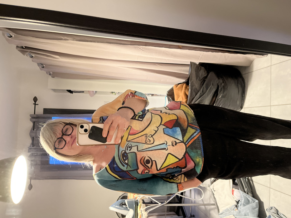 A blonde woman with glasses takes a selfie in a mirror. She wears a Picasso-inspired top and black pants. The background reveals a bedroom with light gray curtains, a lamp, and a black trash bag. An ironing board and clothes are visible in the lower frame. The mirror has a black frame.
