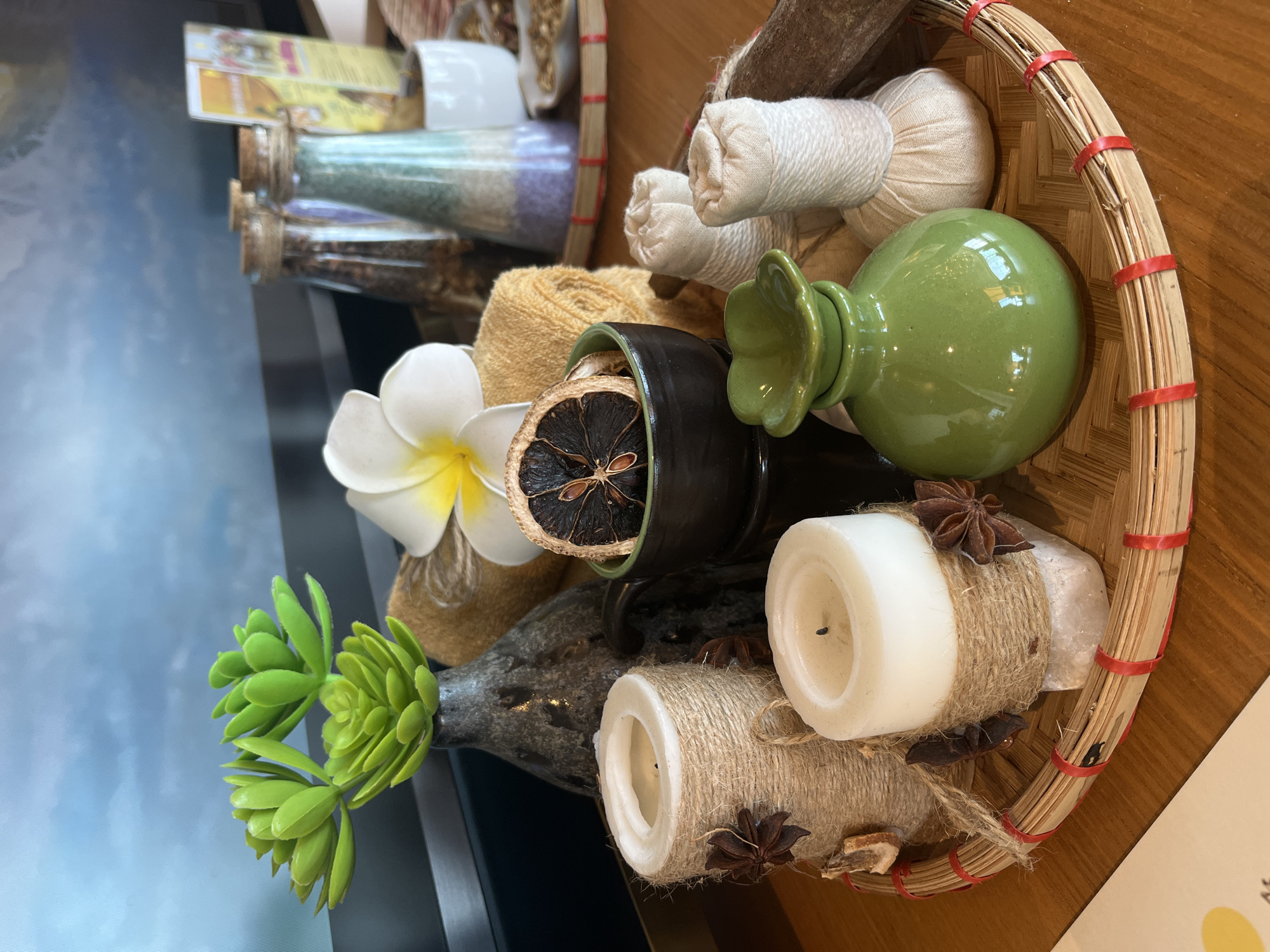 This is a still-life image showcasing a collection of spa and wellness-related items, arranged on a dark surface, likely a table or display shelf. The items are presented in a rustic, natural aesthetic, suggestive of a relaxing and aromatic environment.
In the foreground, a round, woven basket made of light-colored bamboo or rattan with red bindings serves as a central container for many objects. Inside the basket, there are two off-white, cylindrical herbal compress balls, a bright green ceramic pot or diffuser with a fluted top, and two white pillar candles. These candles are wrapped with natural twine and adorned with dried star anise pods. One candle also features a small piece of dried citrus rind.
Outside the basket, to the left, is a white plumeria (frangipani) flower, a dark brown ceramic pot containing a dried, burnt-orange slice of citrus, and what appears to be spools of yellowish-brown natural fiber or twine. Further to the left, a small, vibrant green succulent-like plant is displayed in a dark, mottled vase or bottle.
In the blurred background, several small glass bottles with cork stoppers are visible, containing different colored granular substances, possibly bath salts or dried herbs. Behind these bottles, a stack of brochures or promotional materials with illegible text is partially seen. The background also features a large, blurry area of light blue and white, which could be a reflection on a surface or an out-of-focus background element like a screen.
The overall scene evokes a sense of tranquility and natural aromatherapy, commonly found in spa settings, especially those emphasizing traditional therapies. The items, particularly the woven basket, herbal compresses, and plumeria flower, align with the aesthetic often associated with wellness practices in Southeast Asian locations like Đà Nẵng, Vietnam. No direct human activity is depicted. The lighting is even, suggesting an indoor setting without specific time-of-day cues.