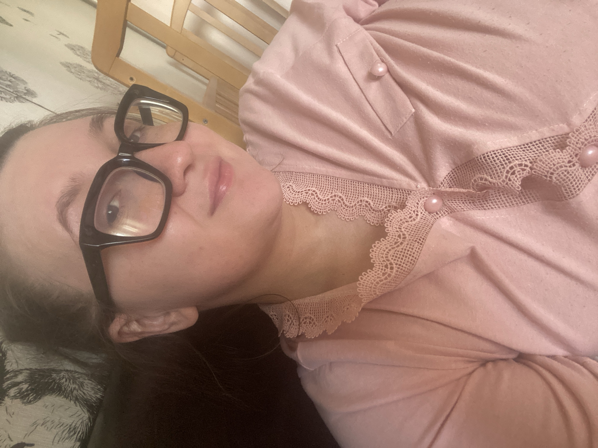 An indoor portrait shot features a female individual of fair complexion, observed from the chest up, situated in Chaikovsky, Russia. She wears dark-framed eyeglasses and a light pink garment, possibly a casual top or pajama shirt, detailed with a V-neckline featuring a light pink lace trim. Similar lace and three spherical light pink buttons extend down the front opening, and a small flap with another matching button is present on the upper right chest area. Her dark hair is pulled back, with a few strands visible near her face. She exhibits a subtle smile with visible lips that appear glossy. The background on the left consists of a dark, possibly upholstered surface, topped by a light-colored wall with a faint, dark, patterned design. To the right, a light-colored wooden slatted structure, potentially part of a crib or bed frame, is visible. The scene is illuminated by soft, indirect light.