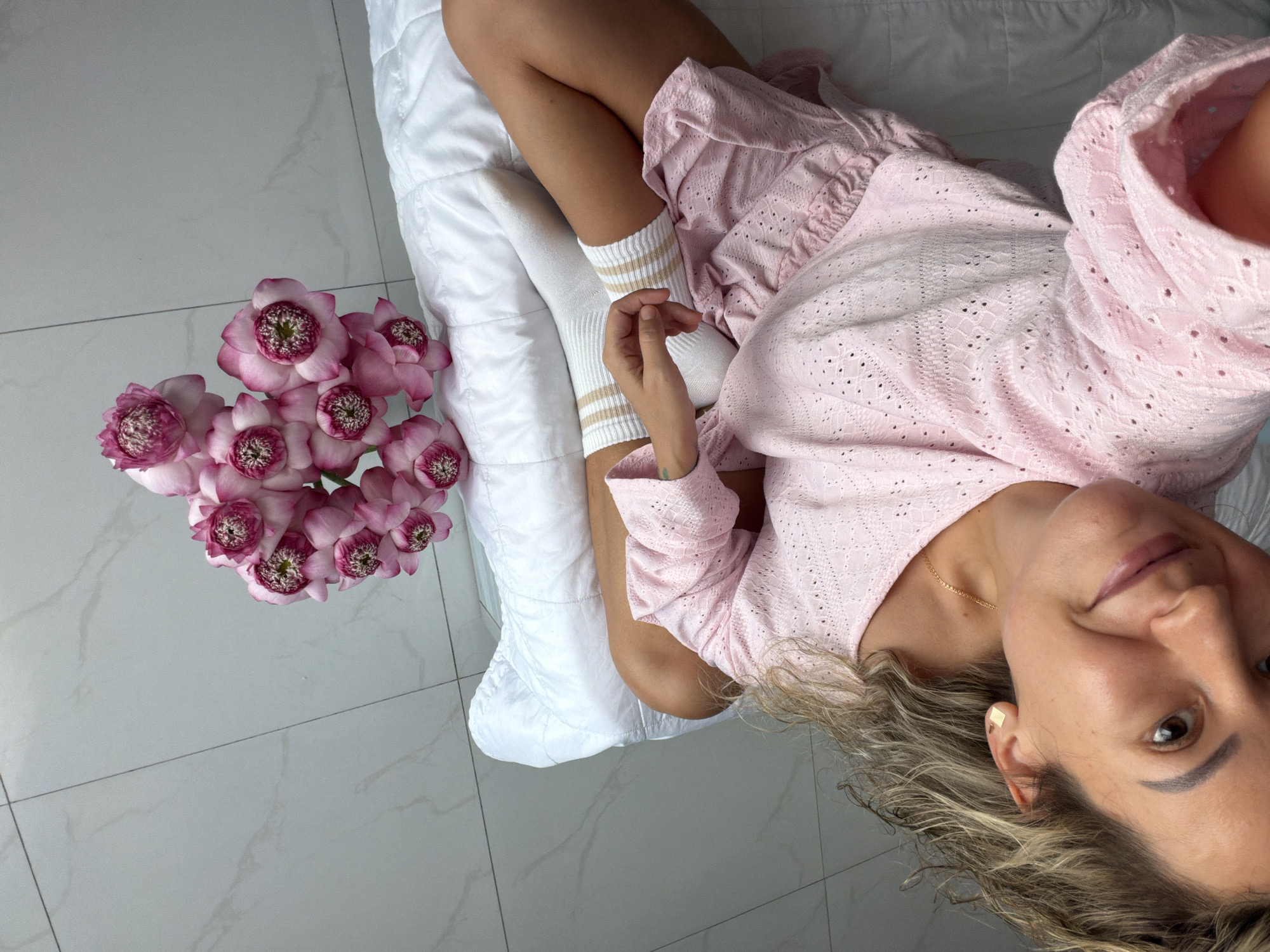 This is a top-down, selfie-style photograph taken from above. The main subject is a woman lying on a white comforter. She has blonde, wavy hair and is wearing a light pink, eyelet lace top and white socks with beige stripes. Her face is tilted towards the camera, and she is smiling, conveying a relaxed and content emotion.
To the left of the woman, there is a bouquet of pink flowers with intricate centers, possibly lotus flowers. The background is a light-colored tiled floor, suggesting an indoor setting. The lighting appears bright and natural, hinting at daytime. The overall scene evokes a sense of domestic comfort and leisure.
The image lacks any visible text. Based on the provided location context of Nha Trang Ward, Vietnam, the setting could be a bedroom or a living area within a home or hotel in that region. The weather is not explicitly shown, but the bright lighting suggests it is likely clear or partly cloudy.