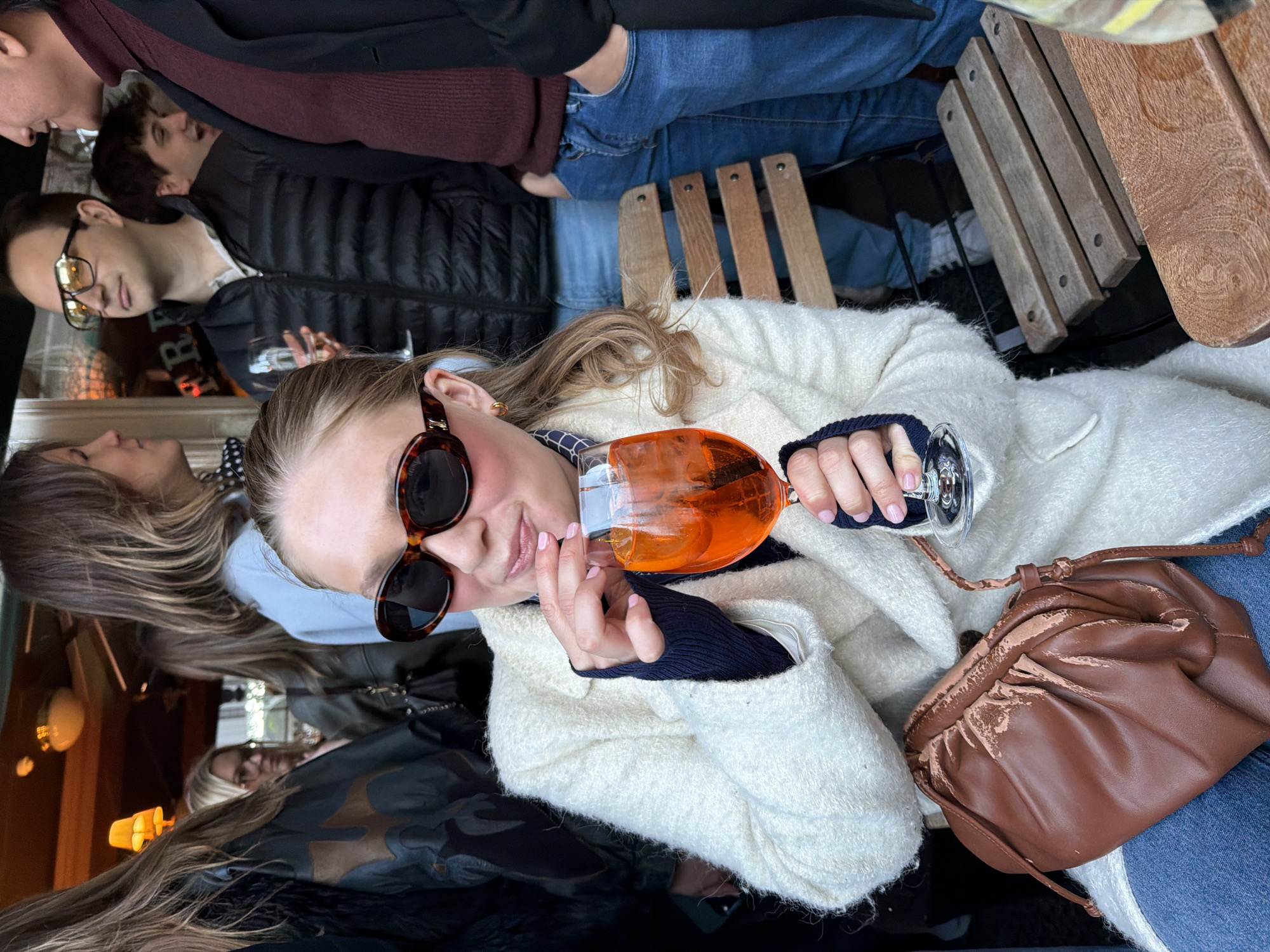 Here is a detailed description of the image:
**Scene**: The image depicts a social gathering at an outdoor seating area, likely a restaurant or bar, in London, United Kingdom.
**People**:
* A woman, the primary subject, is seated. She wears sunglasses, a cream-colored, textured jacket, and a dark top underneath. She holds a glass of an orange-colored beverage.
* Several other individuals are partially visible, including men and women. Some are standing, and others appear to be seated.
**Objects**:
* A wine glass filled with an orange-colored drink, possibly an Aperol Spritz.
* A brown, gathered leather handbag sits next to the woman.
* Wooden chairs and tables indicate an outdoor setting.
* Other glassware can be seen among the other people.
**Actions and Interactions**:
* The woman is holding her drink and looking toward the camera.
* The presence of others suggests a social gathering with interactions among the people present.
**Environment**:
* The setting is outdoors, with a wooden seating area.
* There are other individuals present, suggesting a bustling environment.
* The presence of glassware and furniture indicates a restaurant or bar setting.
* The overall scene suggests a relaxed social gathering.