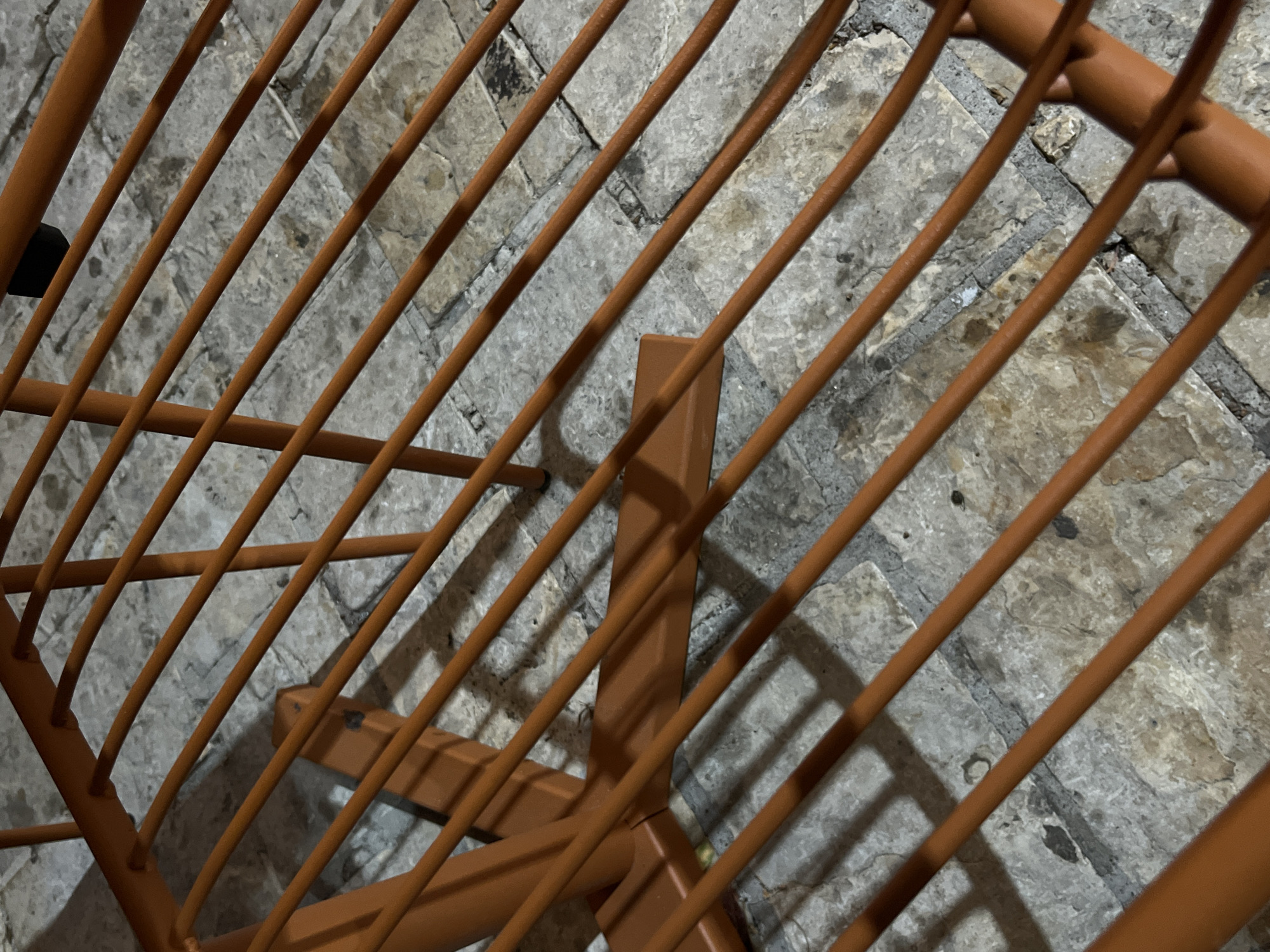 The image shows a close-up of an orange metal chair or table with parallel bars, casting shadows on a stone-tiled surface. The location is Budva, Montenegro. The scene is outdoors, possibly a patio or promenade, given the stone paving and natural lighting. There are no people or animals visible in the frame. The object appears to be made of painted metal tubing and is positioned in a way that suggests it is part of outdoor furniture. The stone tiles show variations in color and texture, indicating they are likely weathered. The overall impression is one of an unoccupied outdoor setting in a Mediterranean city.