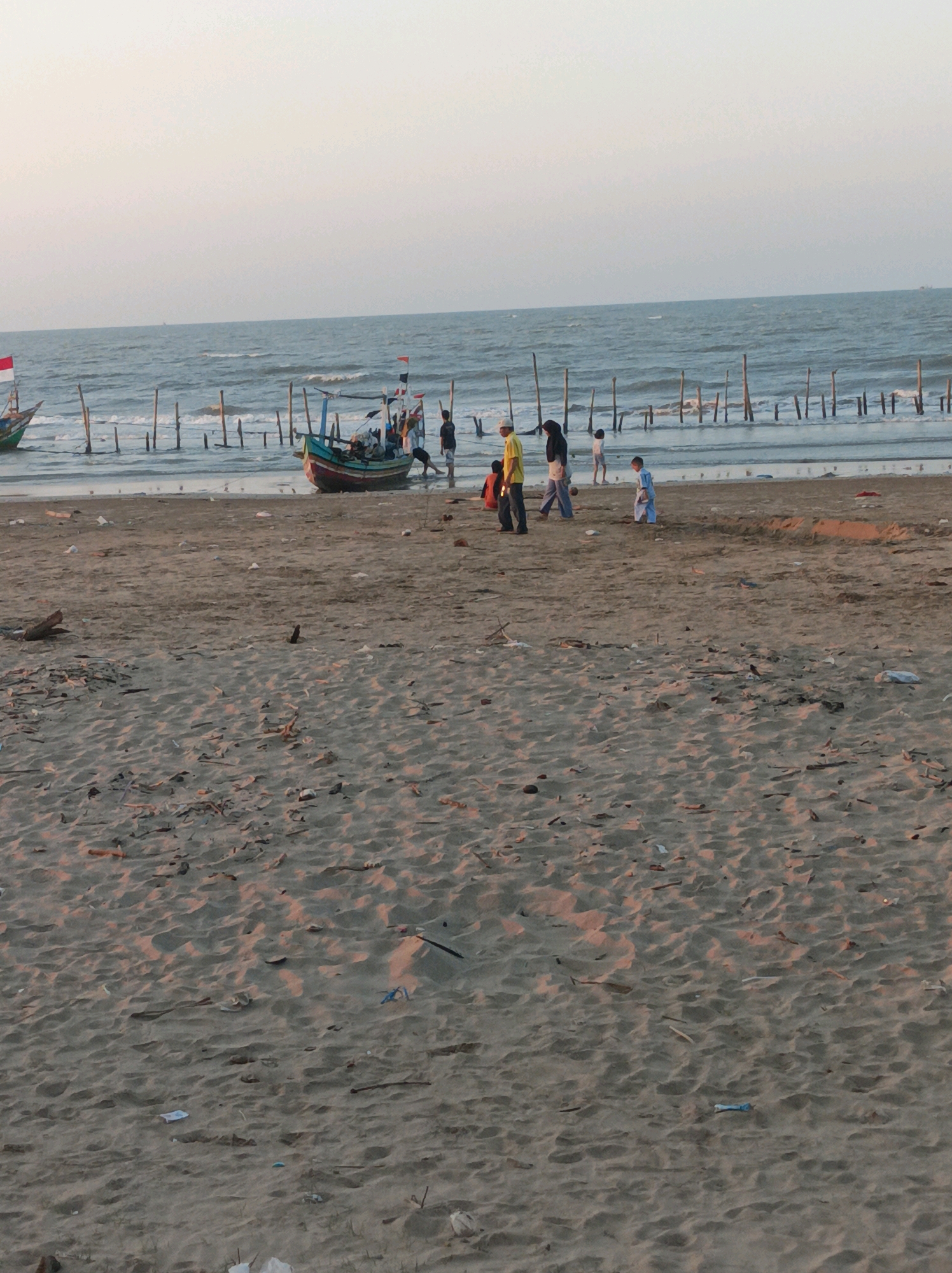 The image depicts a beach scene in Palang, Indonesia. In the background, a body of water, likely the sea, is visible with gentle waves. Several wooden posts are planted in the water, forming a barrier or a structure. Two boats are present, one in the foreground partially submerged in shallow water, and another further back. Several people, including adults and children, are scattered across the sandy beach. Some are standing near the boats, while others are walking or playing. The beach itself is covered in sand with debris, including sticks and plastic waste, scattered across its surface. The sky is a pale, overcast hue, suggesting either early morning or late afternoon light.