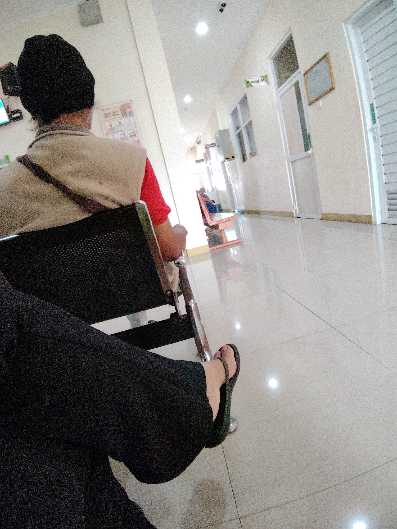 The image shows a dimly lit hospital hallway in Semarang, Indonesia. In the foreground, a person wearing a black beanie, a light-colored vest over a red shirt, and dark pants is seated in a black mesh chair. Their legs are crossed, and they are wearing dark flip-flops. To the left, another person's legs, clad in black pants and wearing dark flip-flops, are visible, suggesting they are seated in front of the person in the chair. The hallway features cream-colored walls, a polished tiled floor reflecting the overhead fluorescent lights, and several doorways. One doorway, marked with a green sign reading "Ruang AS," leads into a room. Further down the hallway, a pink wooden bench is visible, occupied by a person wearing a blue shirt. The environment suggests a waiting area, likely in a public facility.