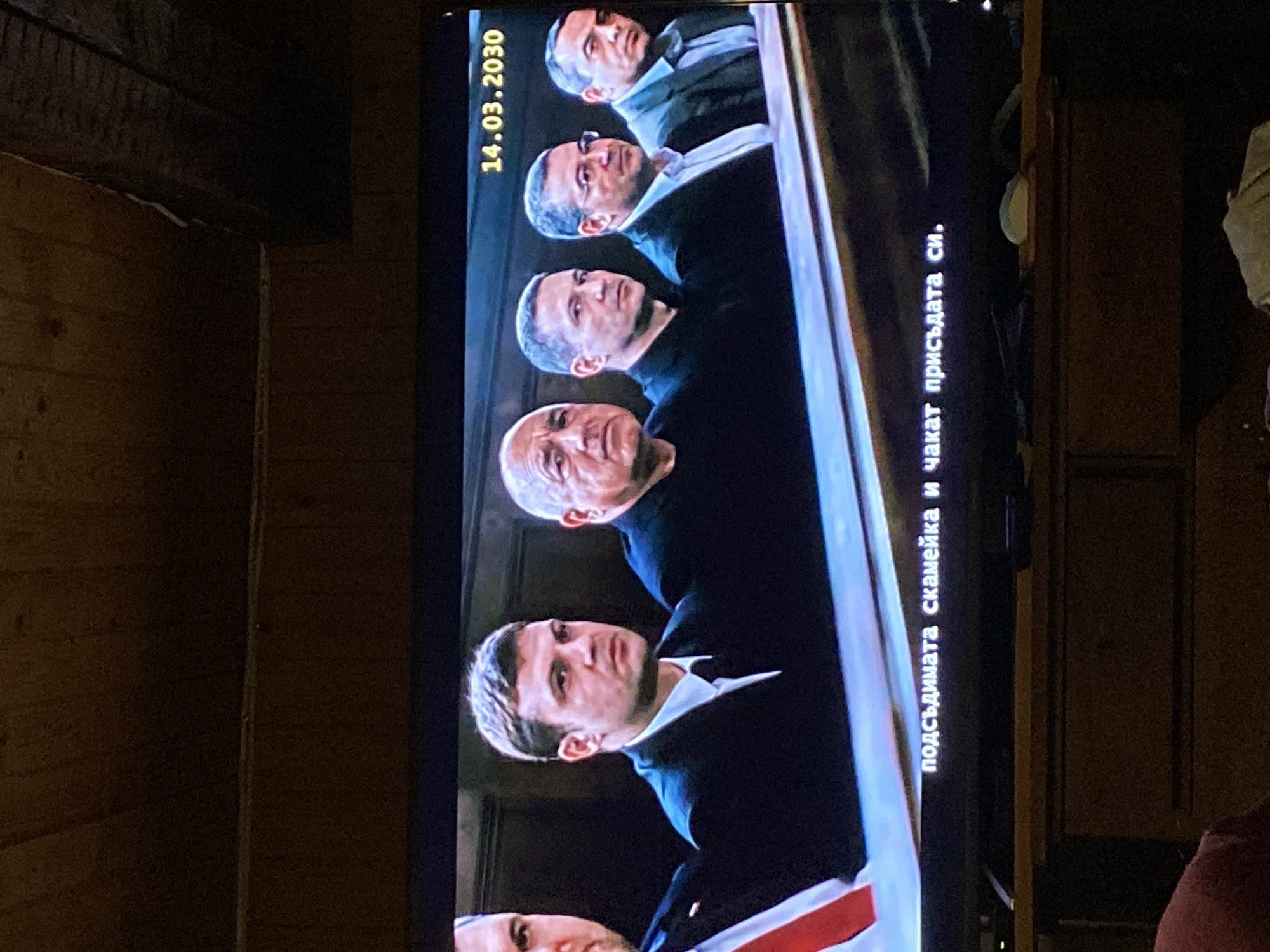 A television screen displays a scene showing five men in suits seated, with the date "14.03.2030" visible at the top left. Subtitles in Bulgarian are present on the right side of the screen, reading "подсъдимата скамейка и чакат присъдата си." (The defendants bench and waiting for their verdict.) The television is set in an indoor environment. The background consists of wooden paneling on one side and a cabinet on the other.