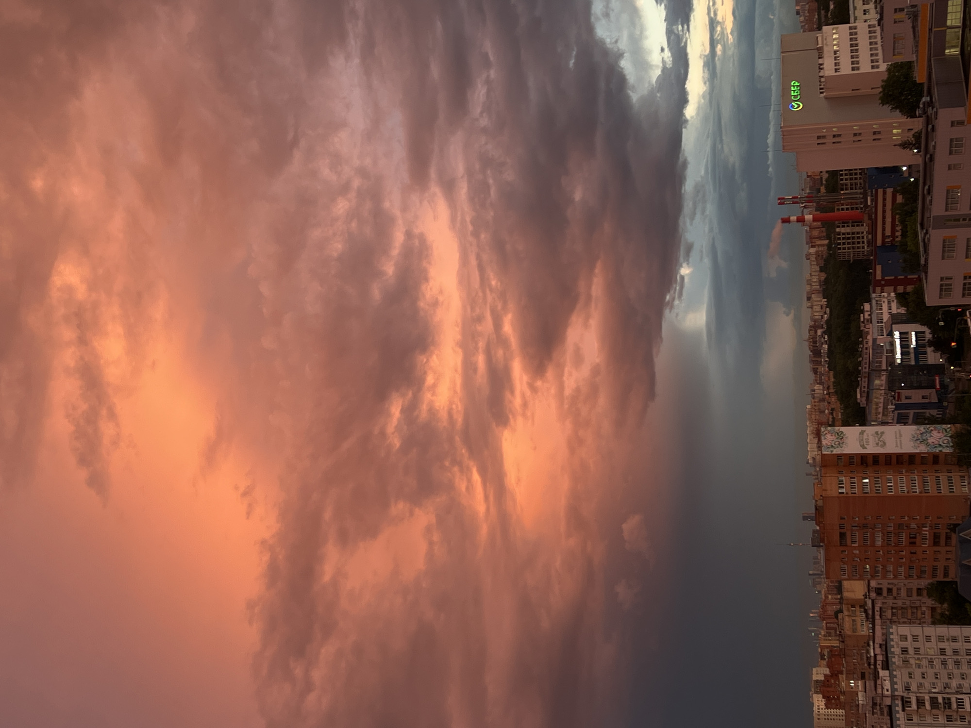 The image shows a wide cityscape at sunset, with a dramatic sky filled with a mix of orange, pink, and dark gray clouds. In the foreground, various residential buildings of different heights and architectural styles are visible, some appearing older and others more modern. Some buildings have visible windows and balconies. In the mid-ground, there are industrial elements including two tall, red and white smokestacks emitting smoke, and a larger white building with a green logo that appears to say "СБЕР". In the background, a dense urban landscape stretches to the horizon, with more buildings and a tall tower visible in the distance. The overall scene depicts an urban environment under a striking, colorful sky. The location is identified as Mytishchi, Russia. There are no people or discernible actions captured in the image.