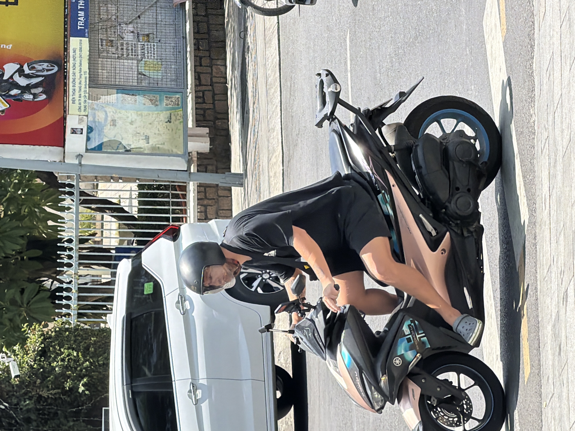 A man wearing a black helmet and a black t-shirt with black shorts is riding a dark-colored scooter on a street in Nha Trang, Vietnam. The scooter has a light brown and dark blue design on its body. He is wearing grey Crocs. In the background, a white SUV is parked behind a white metal fence, and a billboard with a picture of a motorcycle is visible. The street has yellow and white road markings. The scene is illuminated by sunlight, casting shadows on the road.