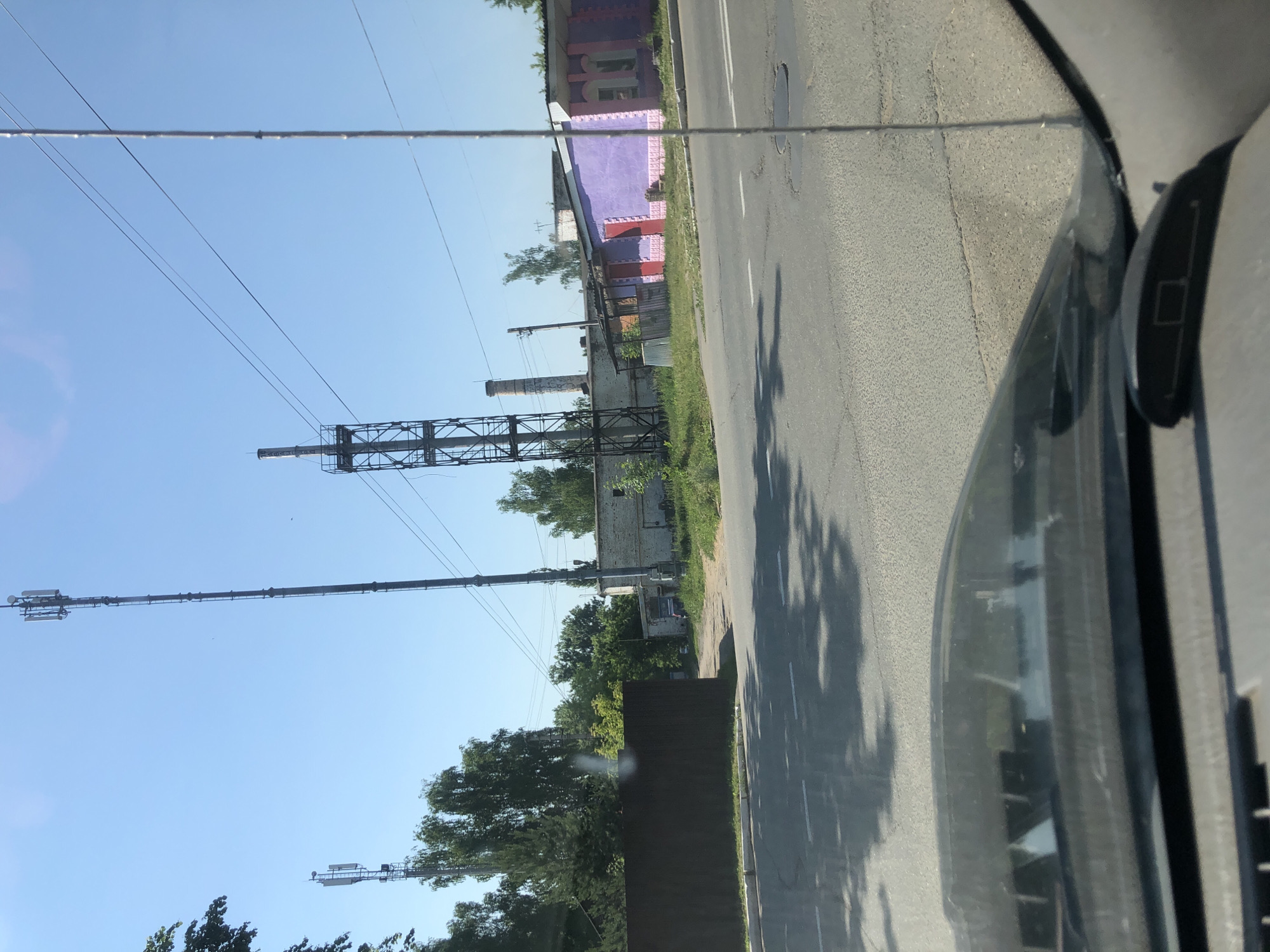 The image shows a view from inside a vehicle, looking out at a street scene with industrial and commercial buildings under a clear blue sky. Two tall, lattice cell towers are visible in the background, one on the left and a taller one on the right. Between them, a shorter industrial chimney with a metal structure around its base is also present. To the right of the chimney, a low-rise building with a predominantly purple facade and pink trim is visible, with red doors. Further to the right, a light-colored brick building with a gray roof is seen. In the foreground, the hood and windshield of the vehicle are visible, with reflections of trees and the sky on the windshield. A dark brown fence runs along the left side of the road. The road itself appears to be asphalt with white lane markings and some potholes. The overall environment suggests an urban or semi-industrial area. This scene is located in Maksimovo, Russia.