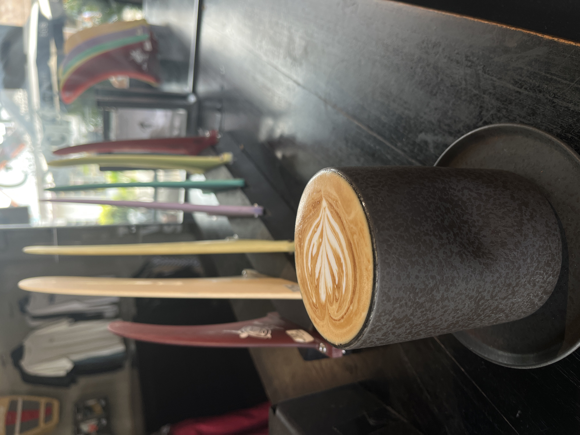 A close-up, eye-level shot shows a dark, textured ceramic mug filled with a latte, artfully decorated with a rosetta pattern in the foam. The mug sits on a matching dark saucer, which rests on a dark, distressed wooden table. In the background, several colorful surfboard fins are displayed vertically on a shelf, along with some t-shirts hanging on a rack. The scene appears to be inside a shop in North Kuta, Indonesia, likely a surf shop or a cafe that caters to surfers, given the prominent display of surfboard fins and apparel. The lighting is somewhat dim, with natural light filtering in from a window or overhead source, creating a soft focus effect on the background elements.