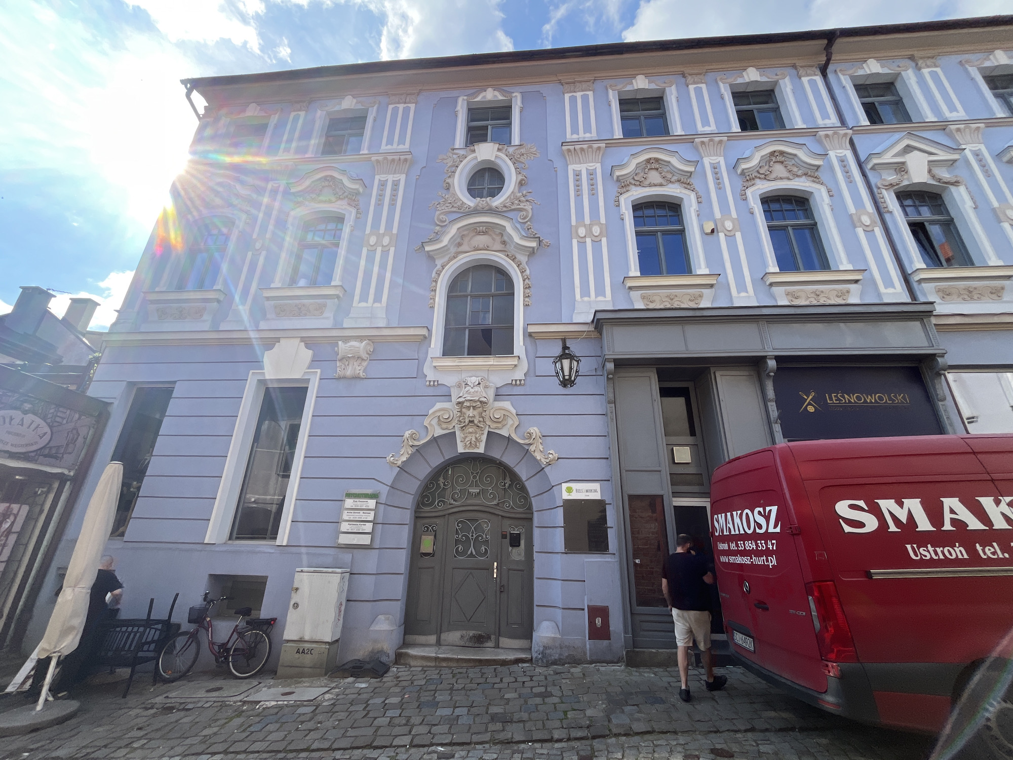 The image captures a street scene with a pastel blue building facade featuring ornate architectural details and arched windows. Sunlight creates a lens flare effect across the upper left portion of the image. A red van with "SMAKOSZ" branding is parked in front of a shop. Two men are visible in the scene; one is standing near the red van, and the other is walking away from the camera. A bicycle and outdoor furniture are present on the sidewalk. The ground is paved with cobblestones. A sign above a doorway on the building reads "LEŚNOWOLSKI". The overall impression is of a European urban setting.