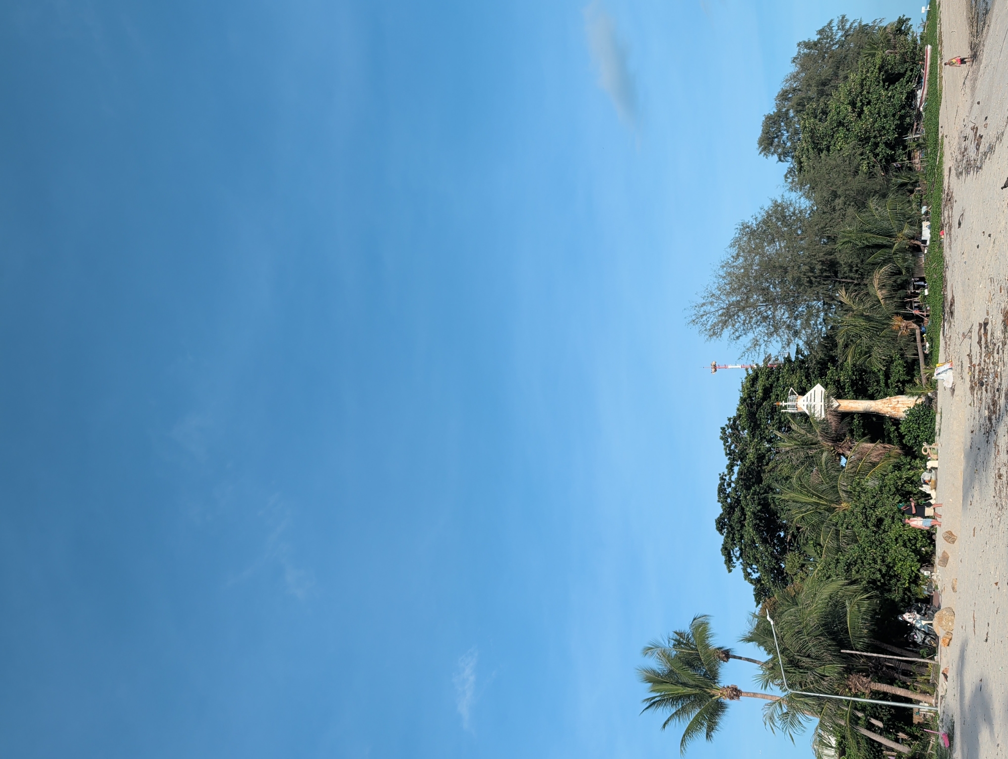 The image depicts a beach scene in ตำบลเกาะพะงัน, Thailand. A clear, blue sky dominates the left side of the frame. The right side shows the sandy beach with vegetation, including palm trees and denser foliage. A white structure resembling a small watchtower is visible amidst the trees. A few individuals are present on the beach, appearing to be walking. Debris and litter are scattered along the shoreline.