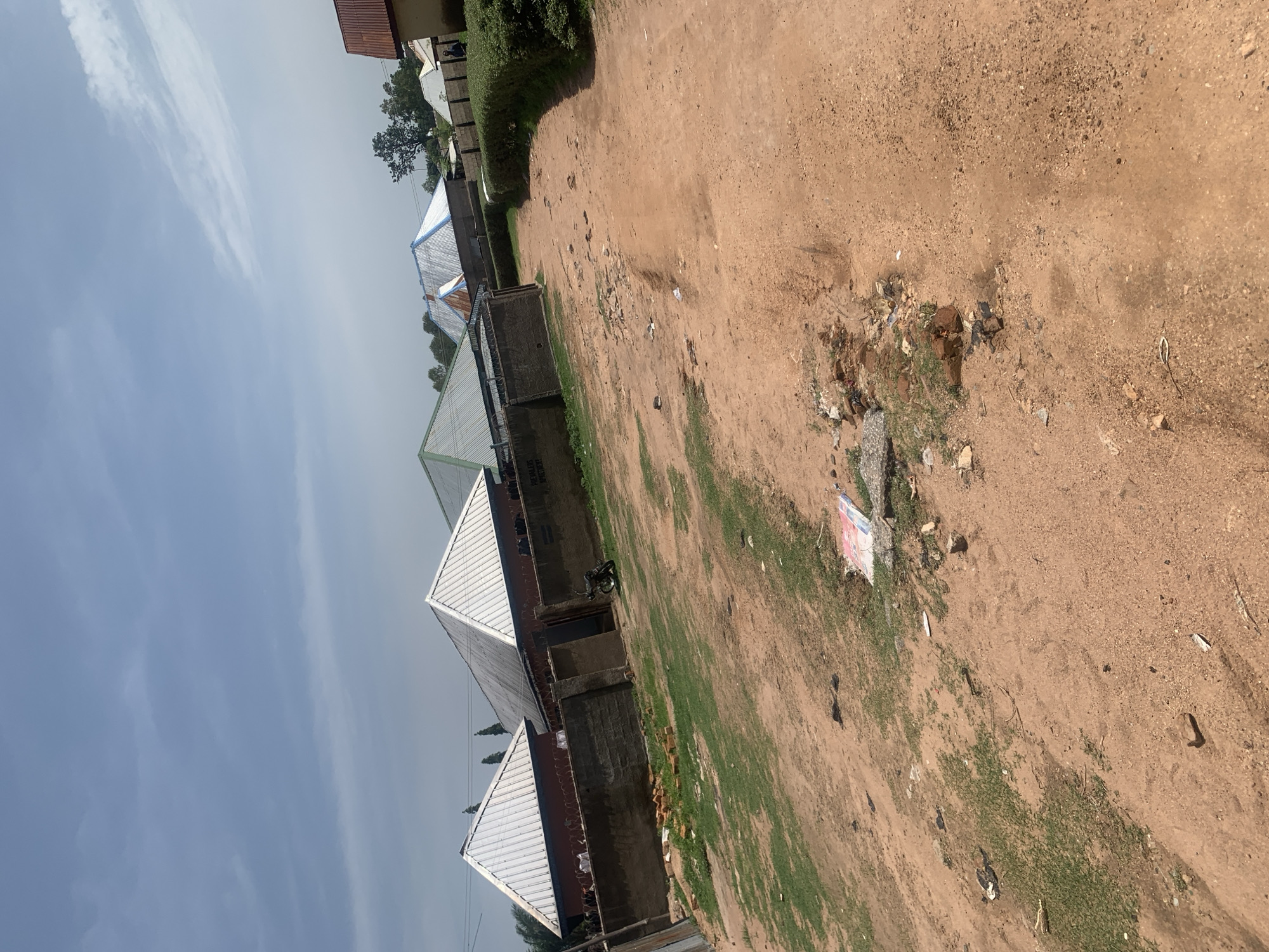 The image shows a rural street scene in Jauro Yinu, Nigeria. The foreground is a dusty, reddish-brown dirt road with patches of sparse green grass. Debris, including bricks, stones, and scattered paper, litters the roadside.
In the middle ground, there are several residential buildings with corrugated metal roofs, some appearing silver and others showing some discoloration. These houses are enclosed by concrete block walls topped with barbed wire, suggesting security measures. A motorcycle is parked near an entrance to one of the compounds.
The background features more trees and vegetation, with the sky above being mostly clear blue with some wispy white clouds. The overall impression is of a modest, somewhat unkempt residential area under a bright sky.