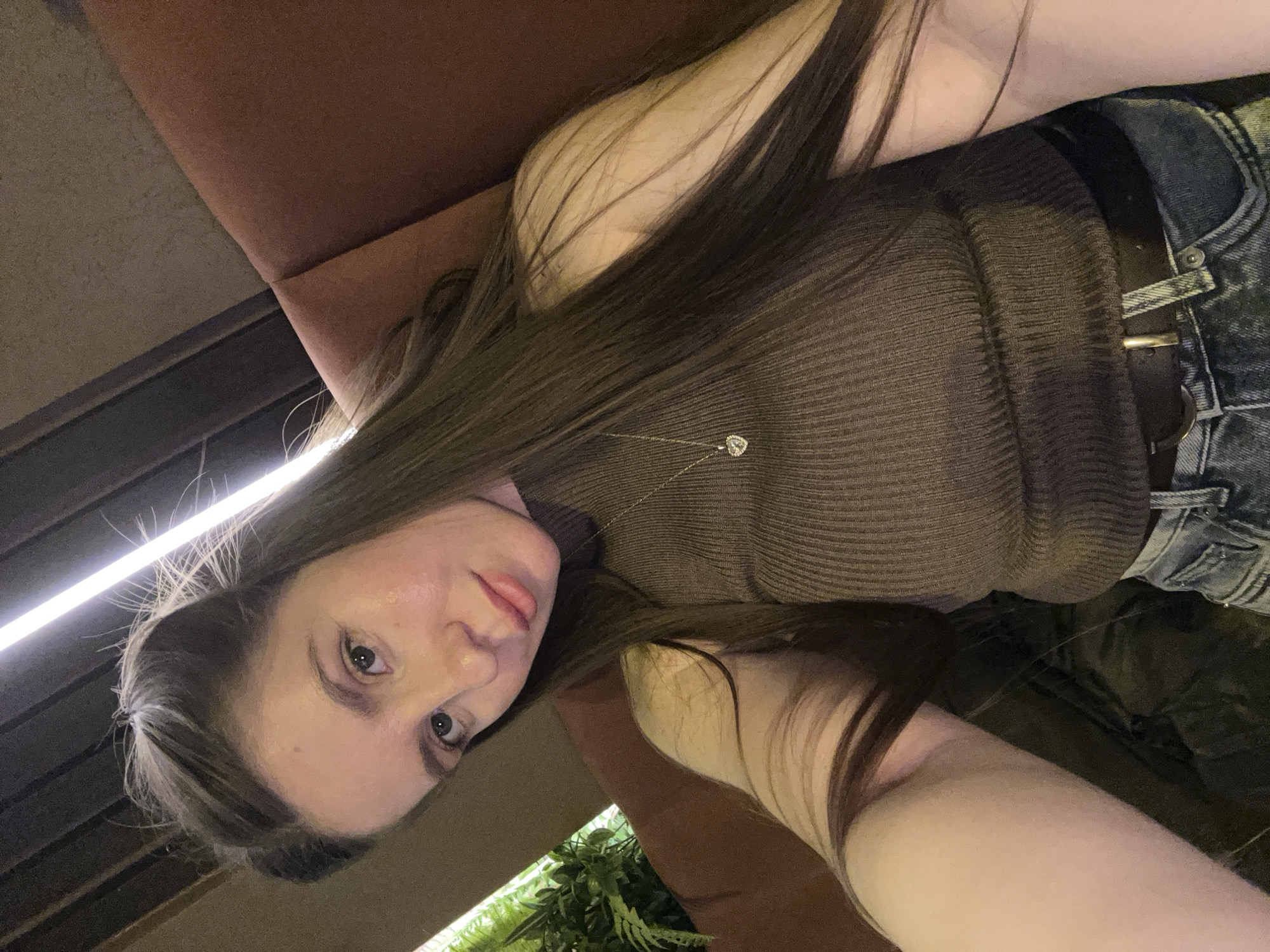 A young woman is depicted in a close-up, slightly tilted portrait, appearing to be a selfie taken indoors, possibly in a dimly lit restaurant, bar, or lounge in Moscow, Russia. She has long, straight, dark brown hair that cascades over her shoulder and arm. Her head is tilted, and she looks directly into the camera with a neutral to slightly serious expression.
She is wearing a form-fitting, dark brown, ribbed long-sleeved top, paired with light-washed blue denim jeans and a dark belt with a metallic buckle. A silver-colored chain necklace with a small, heart-shaped pendant is visible against her top.
The background is dark and indistinct, featuring a dark brown or reddish-brown upholstered surface, likely a sofa or chair, behind her. A prominent linear artificial light source, possibly an LED strip, shines brightly in the upper left portion of the image, casting highlights on her hair and face. Further in the background, to the lower right, some green foliage from what appears to be an indoor plant is visible. The overall lighting suggests an evening or nighttime setting. No visible text is present in the image.