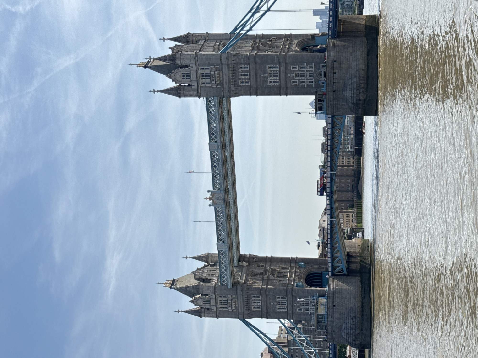 The image showcases the Tower Bridge in London, United Kingdom. The bridge is a prominent architectural structure, featuring two towers connected by a central span. The architectural style is intricate, with detailed stonework and turrets atop each tower. The River Thames flows beneath the bridge, and its waters reflect the bridge's structure and the surrounding urban environment. In the backdrop, buildings and structures of the city are visible. The sky is clear and blue with some clouds. The image appears to have been rotated.