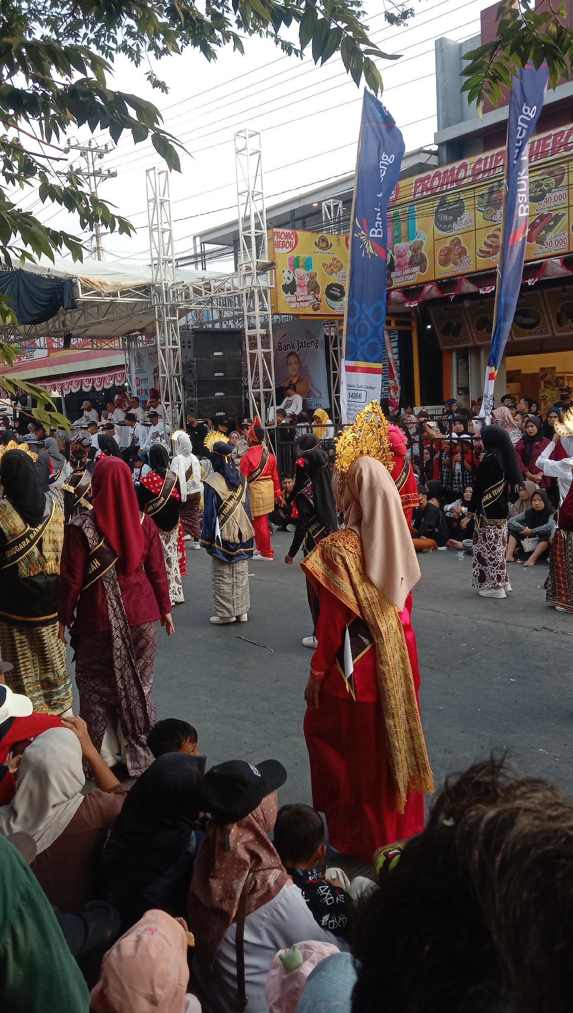 An outdoor event is taking place in Jatisawit, Indonesia. A group of individuals are dressed in traditional attire, participating in what appears to be a performance or parade. They are adorned with colorful fabrics, elaborate headwear, and sashes with inscriptions. Some are wearing red, blue, and maroon outfits, while others have on gold and patterned garments. Several people in the foreground are seated, observing the event, and appear to be a mix of adults and children, some wearing headscarves and hats. In the background, a stage with speakers is visible, along with promotional banners displaying text and images, including a "PROMO HEBOH" sign. The scene is set on a street, with trees and overhead power lines indicating an urban or semi-urban environment.