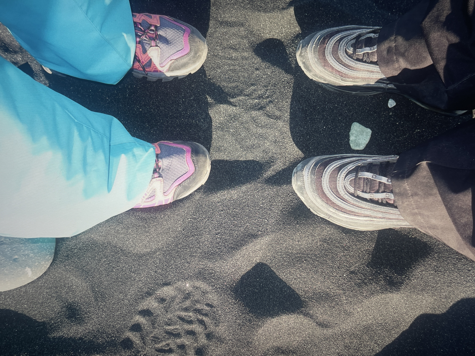 The image shows four pairs of feet standing on black sand. Two pairs of feet wear sneakers with blue and grey pants. The other two pairs of feet wear sneakers with dark pants. There are two small ice chunks on the sand near the dark pants. Footprints are visible in the lower left of the frame. The location is a black sand beach in Iceland.