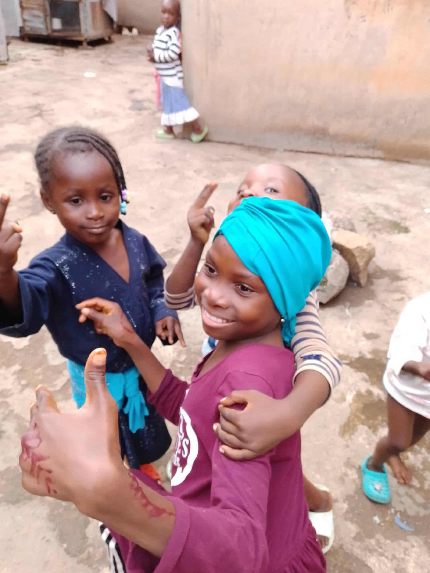 The image depicts a group of young children in what appears to be an outdoor, somewhat unpaved area with a building in the background. In the foreground, a child in a maroon shirt with a white design is giving a thumbs-up gesture, their hand decorated with what looks like henna. This child is wearing a teal headscarf. To their left and slightly behind, another child in a dark blue shirt and bright blue pants is also making a hand gesture, possibly with two fingers raised. Further in the background, a child in a striped shirt and blue and white skirt stands near a wall, wearing light green shoes. Another child in a white shirt is visible on the far right. The ground is made of dirt and scattered stones. The overall scene suggests a casual, everyday moment among children in a community setting.