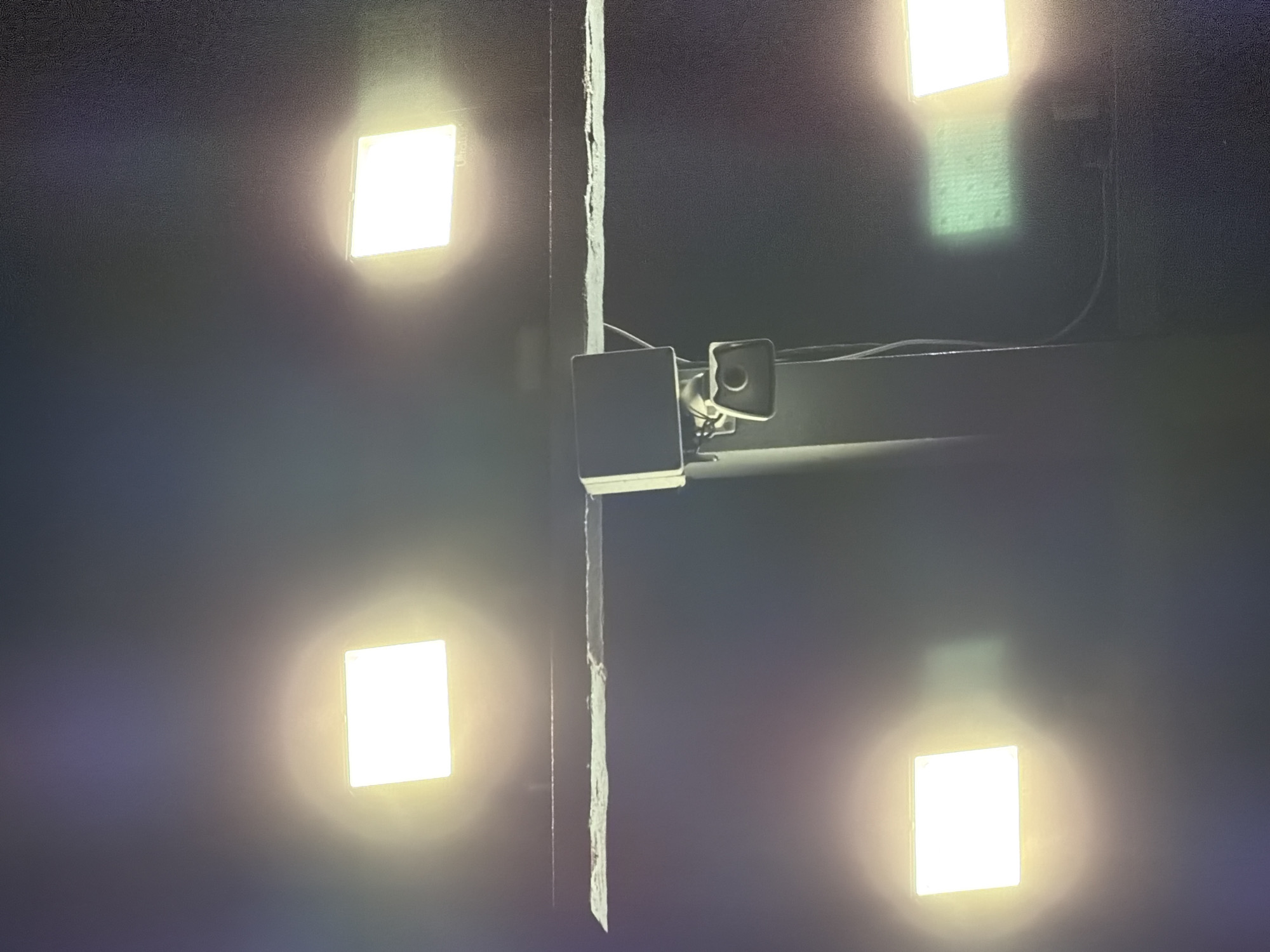 The image shows an exterior scene at night, illuminated by multiple square floodlights. Four prominent floodlights are visible: two in the upper corners, one in the lower left, and one in the lower right. A pole supporting a surveillance camera and a small, dark box is centrally located, extending upwards behind a horizontal beam. The beam has a rough, textured surface with a white stripe running along its length. The background is dark and indistinct, suggesting an outdoor urban environment. The floodlights cast a bright, diffused glow, with some reflecting on unseen surfaces. One floodlight in the lower right appears to be projecting a faint green light onto a textured surface. The overall impression is one of nighttime surveillance or illumination in a public area.