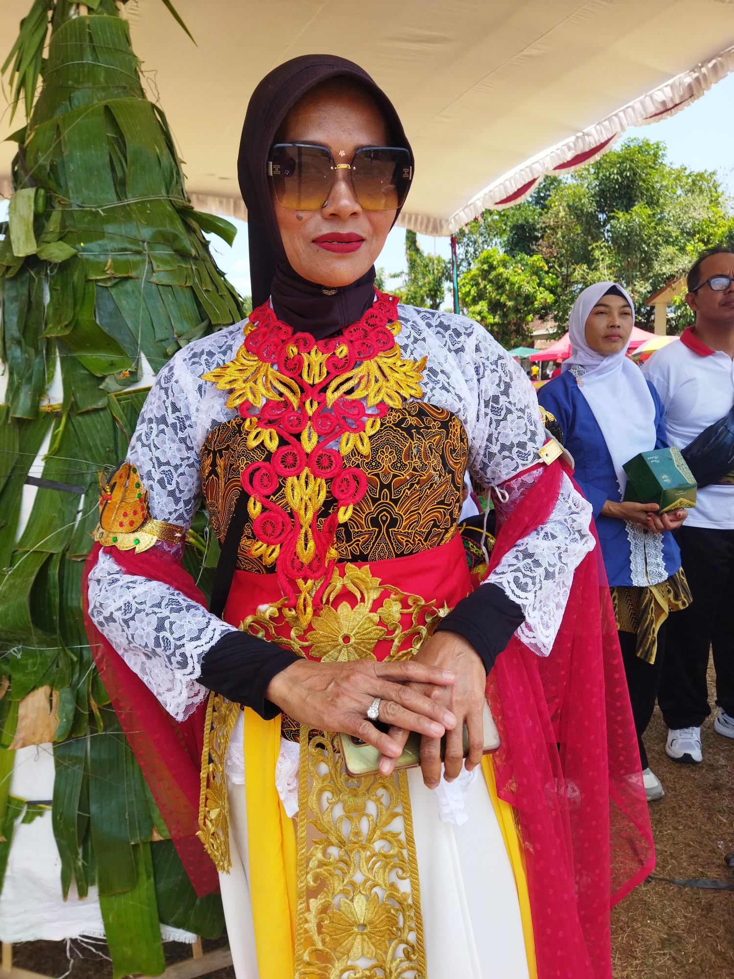 In Semarang, Indonesia, a woman, adorned in elaborate traditional attire featuring white lace, red, gold embroidery, and a dark hijab, stands in front of a conical structure made of large green leaves. She wears oversized sunglasses and red lipstick. Her hands are clasped, one resting on a mobile phone and the other over it, a ring visible on her left ring finger. Behind her, other individuals are present, including a woman in a blue and white headscarf holding a green box, and a man in a white shirt and black pants. The background includes trees, a white canopy with red trim, and a grassy area, suggesting an outdoor event.