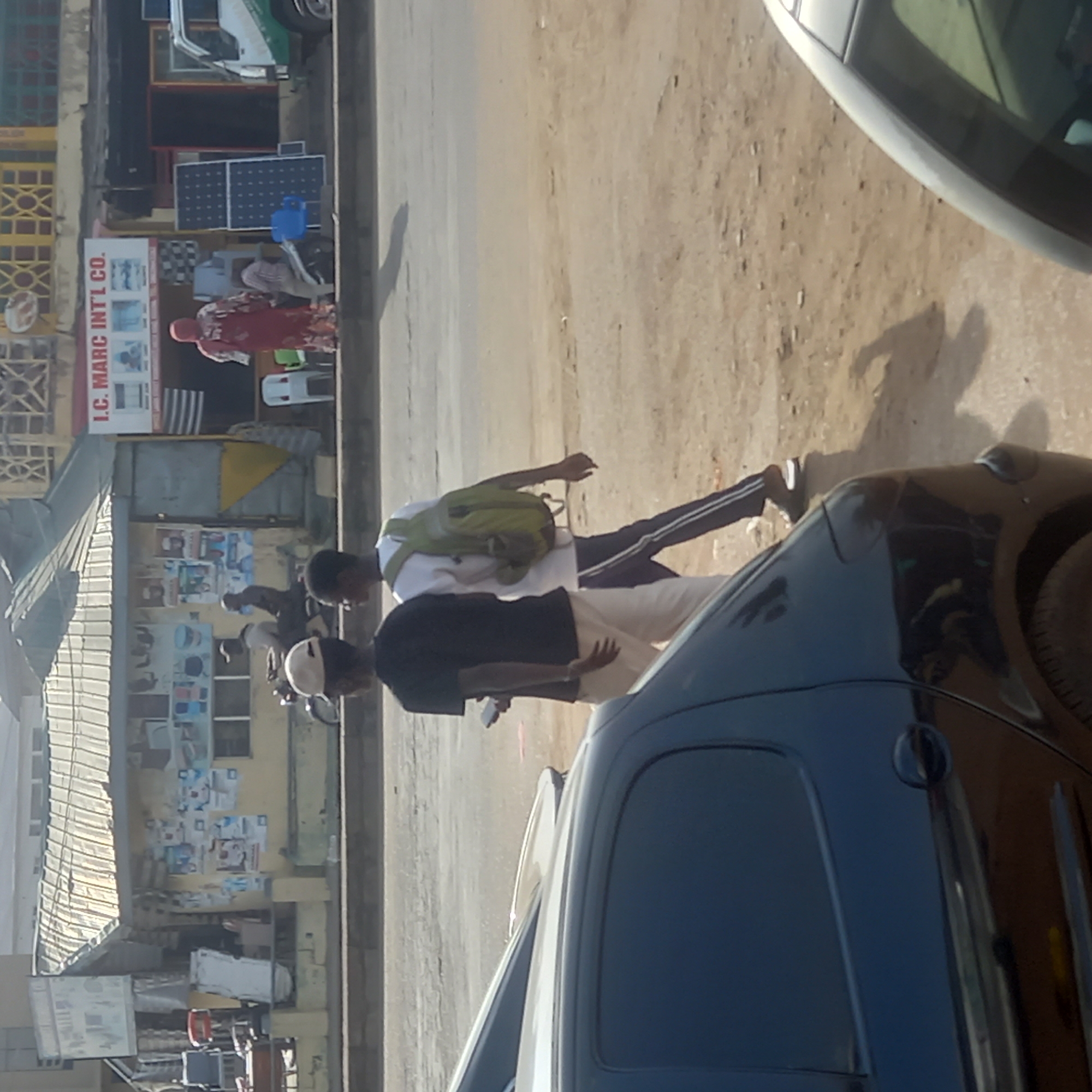 The image shows a street scene in Dadi Riba, Nigeria. Two pedestrians walk along the sidewalk in front of a commercial building. The building has a storefront with advertisements and the business name "I.C. MARC INT'L CO." displayed. Parked cars are visible on the side of the road. One pedestrian wears a black shirt and light-colored pants, and is holding a small object. The other pedestrian wears a white shirt, dark pants with a stripe, and carries a green backpack. The street is paved and appears to be a mix of asphalt and dirt. The weather appears to be sunny.