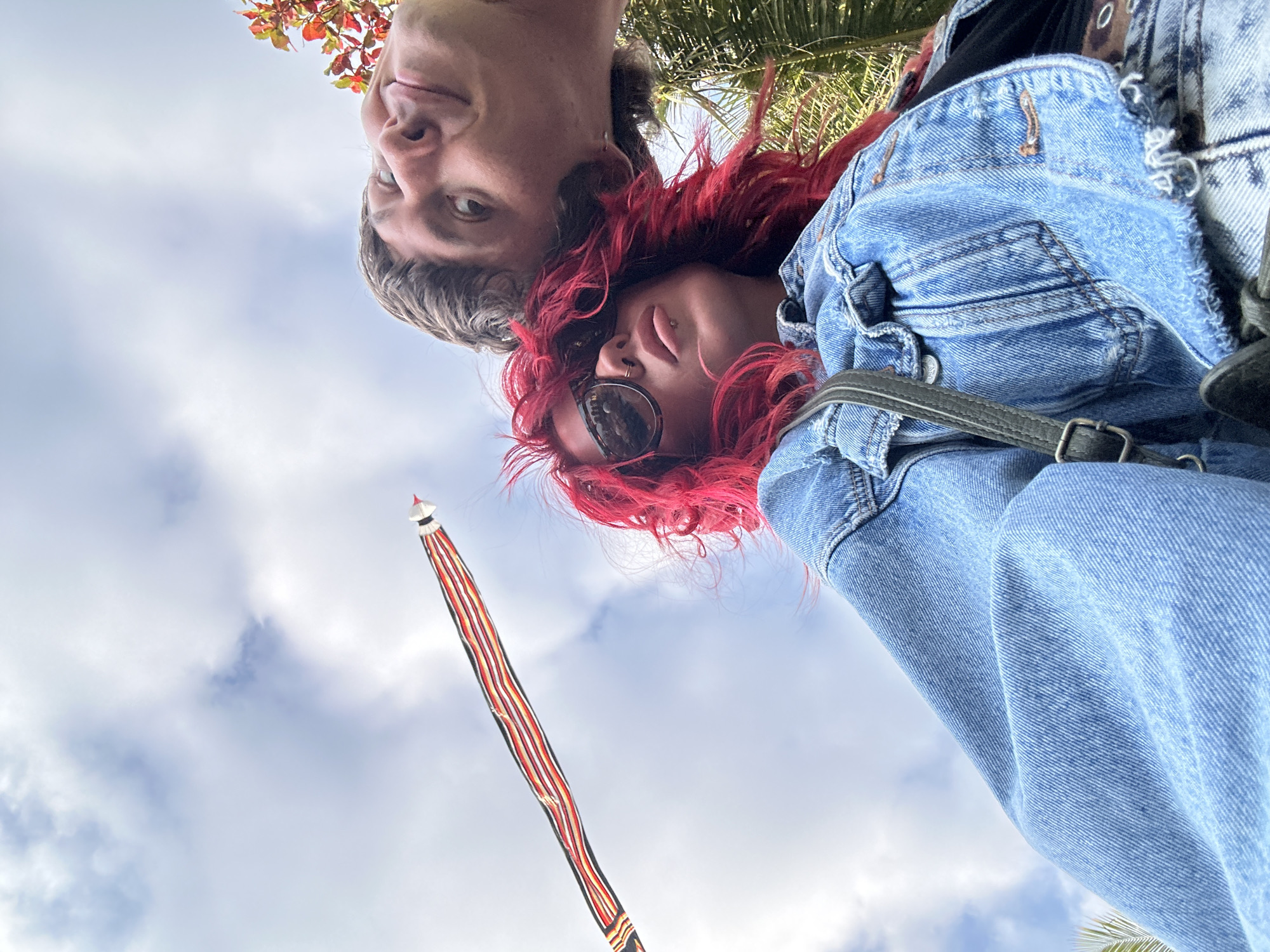 In Mengwi, Indonesia, a low-angle selfie captures two individuals against a backdrop of a cloudy sky and tropical foliage. The person in the foreground, with vibrant red hair and sunglasses, wears a denim jacket and a crossbody bag. To their right and slightly behind, a person with short brown hair is partially visible, their face turned towards the camera. A long, striped kite extends diagonally across the sky from the left.