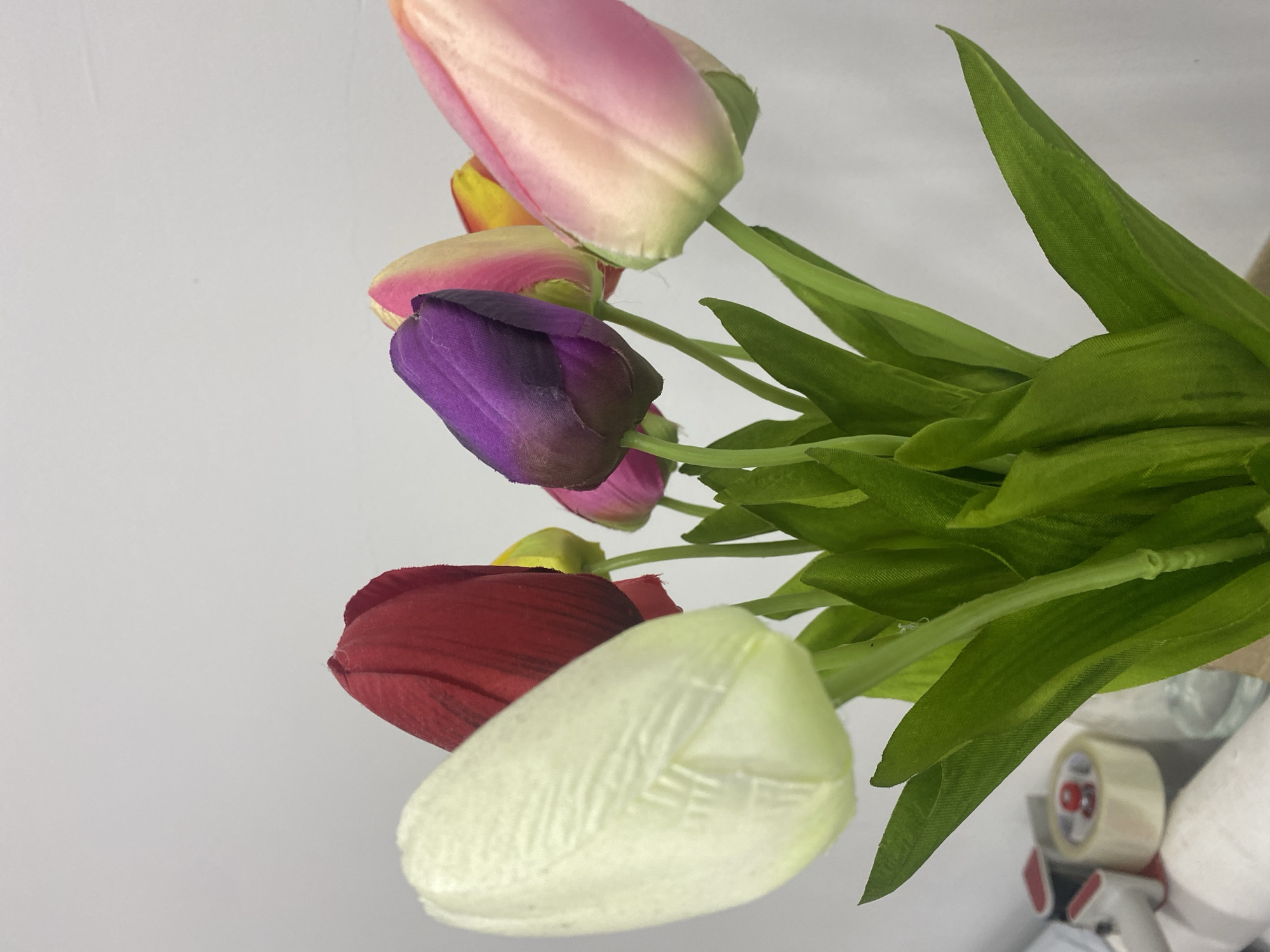 The image displays a close-up, slightly upward-angled view of a bouquet of artificial tulips situated indoors in Chaikovsky, Russia. The foreground features a prominent, out-of-focus white tulip on the left. Behind it, a red tulip is visible, followed by a central purple tulip, and additional pink, yellow, and mixed pink-yellow tulips extending towards the right side of the frame. All tulips exhibit green artificial stems and leaves, with visible fabric textures on the petals, indicating synthetic material. The background consists primarily of a plain light gray or off-white wall. In the lower-left quadrant, several objects are visible out of focus on a surface: a roll of light-colored adhesive tape with a red core, parts of tape dispensers (red and gray elements), a clear container, and a white rolled material. No individuals, actions, or direct interactions are depicted.
