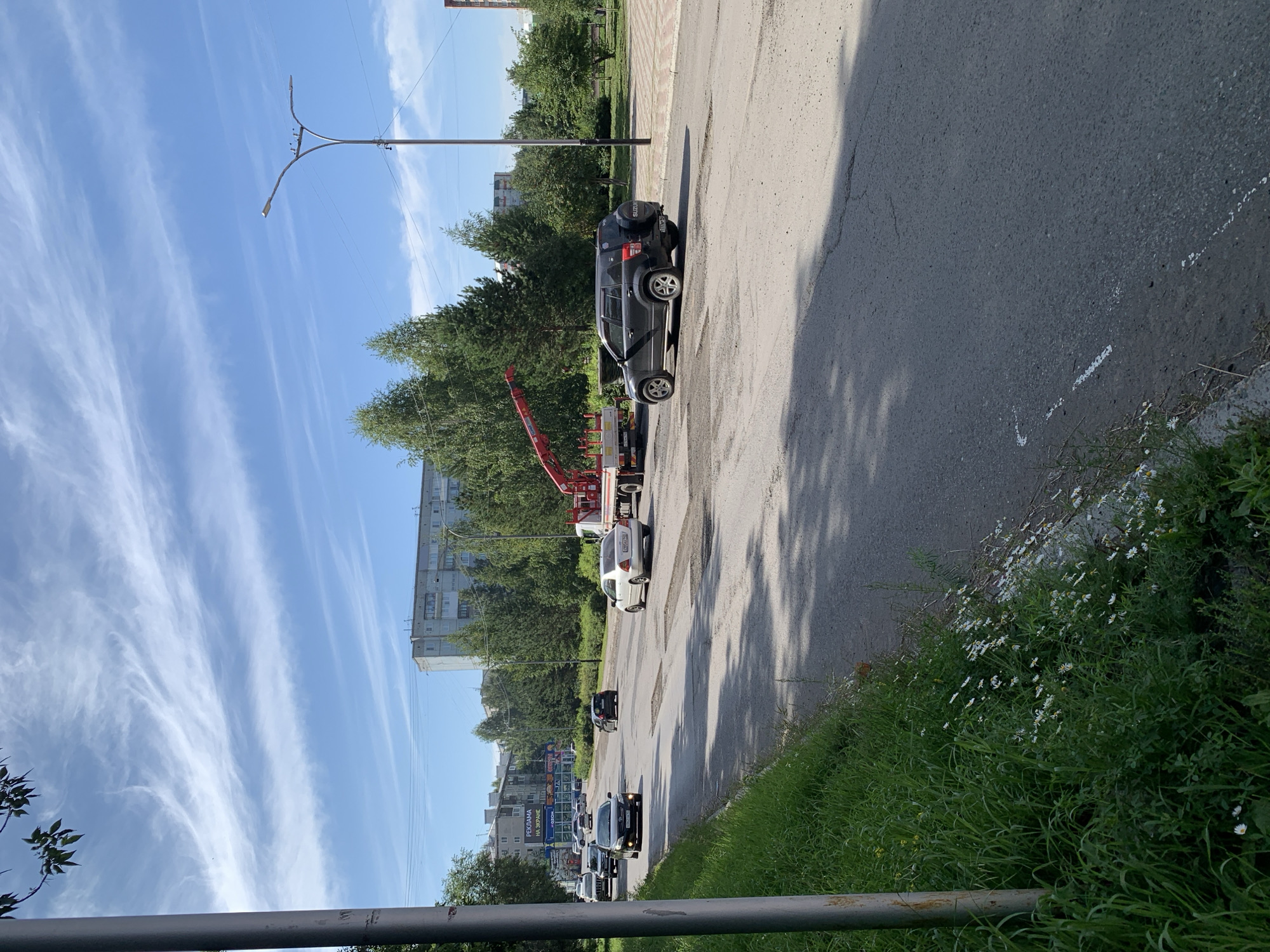 The image depicts a street scene in Prokopyevsk, Russia, on a bright, sunny day with wispy clouds. The road is paved but shows signs of disrepair with several patched areas. Cars are driving on the road, including a black SUV, a white sedan, and several other vehicles further in the distance. A tow truck with a red crane is parked on the right side of the road. On the left side of the road, there is a patch of green grass with small white flowers and a metal pole. In the background, there are multi-story apartment buildings and a line of trees. The overall environment suggests an urban setting within a residential area.