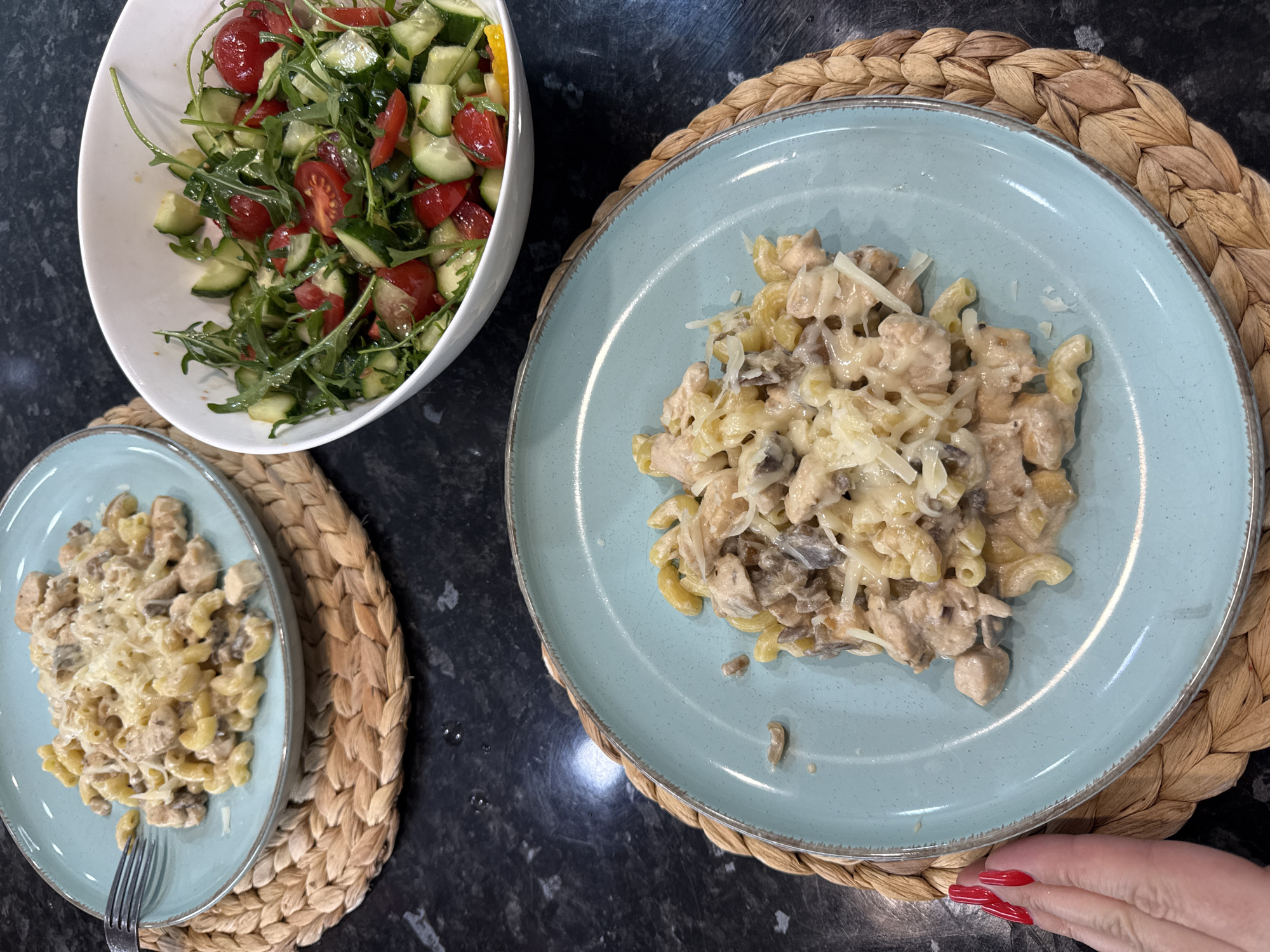 In the city of Sochi, Russia, a close-up, overhead shot showcases a table laden with food. Two light blue plates, each holding a serving of pasta with chicken and mushrooms, are positioned on woven placemats. A fork rests on the left plate, and shredded cheese is sprinkled over the pasta. To the right, a white bowl overflows with a vibrant salad composed of arugula, sliced cucumbers, and cherry tomatoes. The background consists of a dark, speckled countertop. A hand with red nail polish is visible on the left side of the frame, near one of the pasta plates.