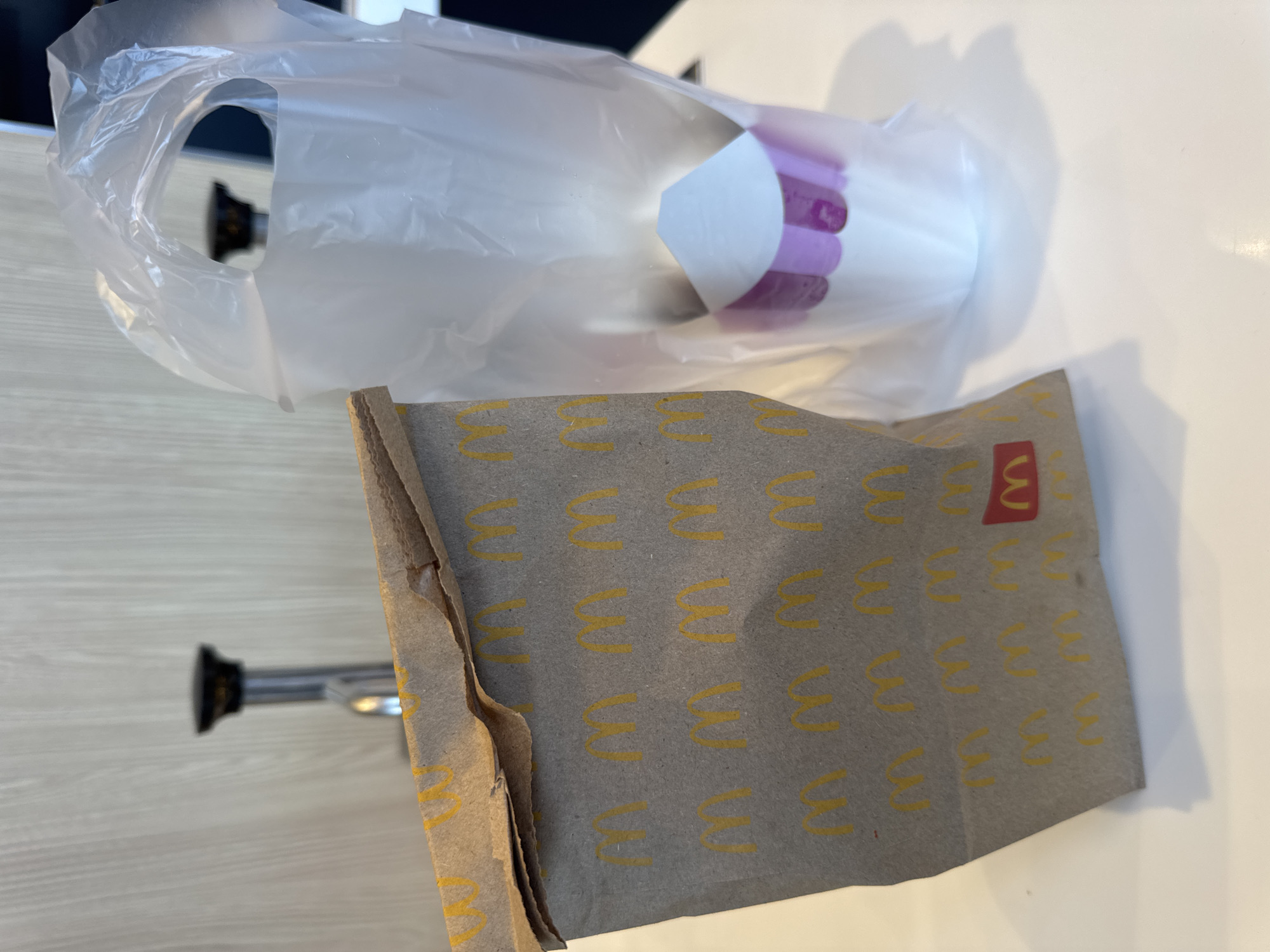 A brown paper bag with repeating yellow McDonald's arches is placed on a white surface next to a clear plastic bag containing a white cup with purple stripes. In the background, a chrome faucet with a black handle is visible against a light wood-paneled wall. The scene is indoors, possibly a fast-food restaurant. There are no people or animals present.