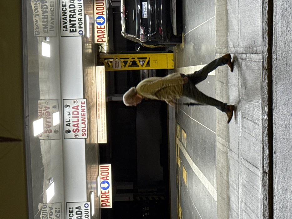 An elderly man with gray hair and wearing brown shoes, green pants, and a beige shirt walks right towards a concrete wall in a parking garage with yellow lines. A black car is parked nearby. Above, there are several signs in Spanish, including "Salida Solamente," "Pare Aqui," "Avance! Entrada por Aqui," and "Aguas." The background is dimly lit.