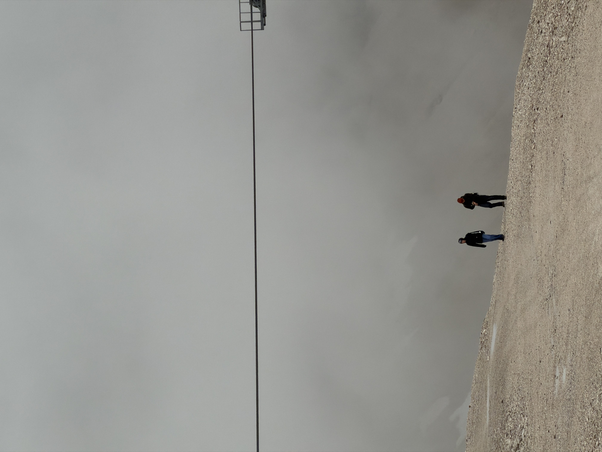 Two individuals are walking on a gravel path on a mountain slope. The sky is overcast and foggy, obscuring the distant landscape. A cable stretches horizontally across the upper portion of the image, possibly part of a ski lift or cable car system. The individual on the left is wearing a black jacket, blue jeans, and a dark beanie, and is walking towards the right. The individual on the right is wearing a dark jacket, dark pants, and a red beanie, and is also walking towards the right. The image depicts a scene at the Zugspitze, Germany's highest peak, located in Garmisch-Partenkirchen.