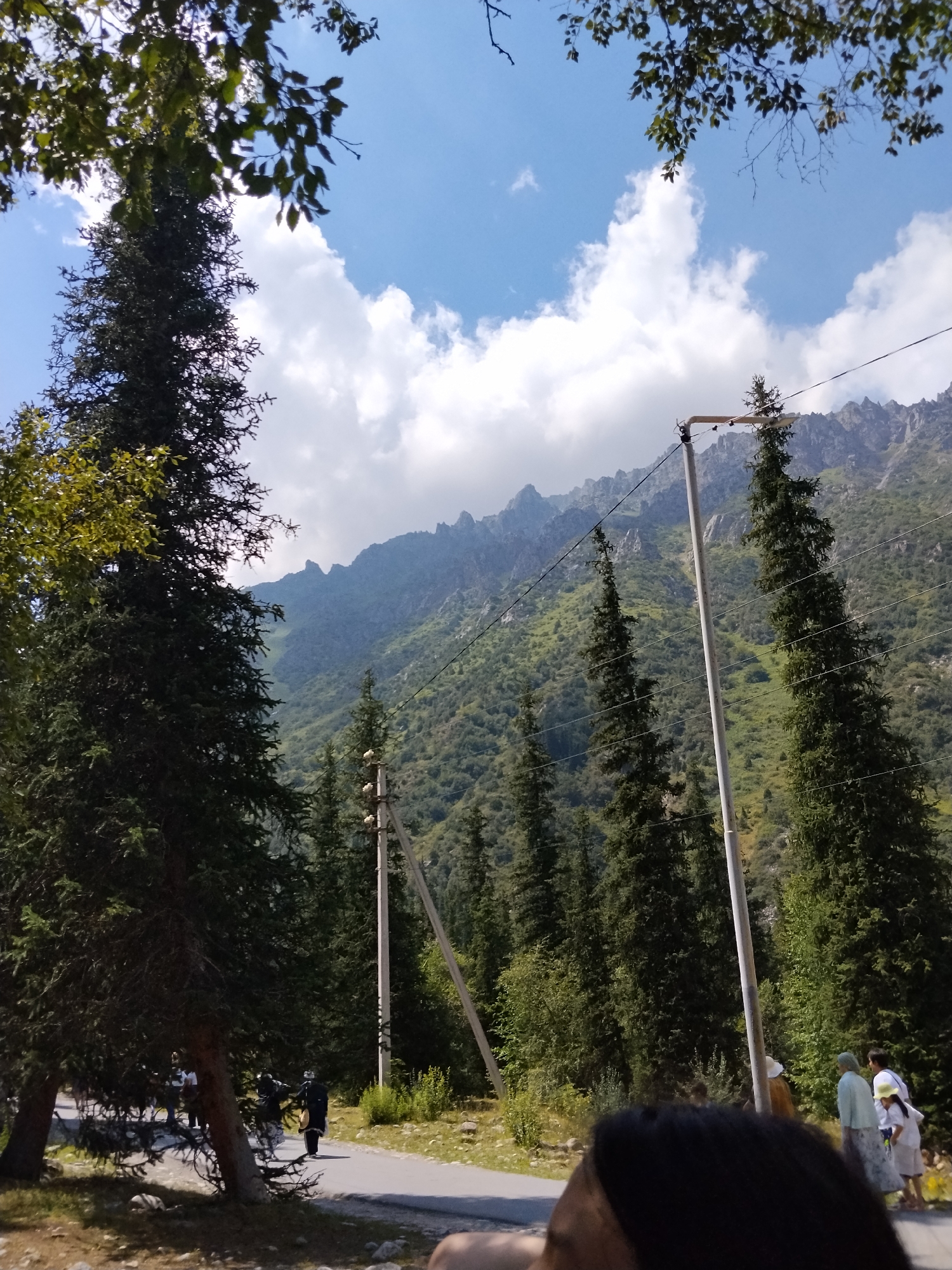The image captures a view of a mountain landscape with coniferous trees and a road. In the foreground, a dark-haired person's head and shoulders are visible, partially obscuring the scene. To the left, a tall evergreen tree dominates the frame. Further along a paved road, several people are walking, some in traditional attire. One person in the foreground appears to be wearing a dark robe and carrying a drum. The road leads towards the mountains in the background, which are covered in lush green vegetation and jagged rocky peaks. Overhead, a blue sky with white clouds provides a backdrop. Power lines and utility poles are also present, suggesting the area is served by electricity. The overall impression is one of a scenic natural environment with human activity. The location is identified as Unknown, Kyrgyzstan.