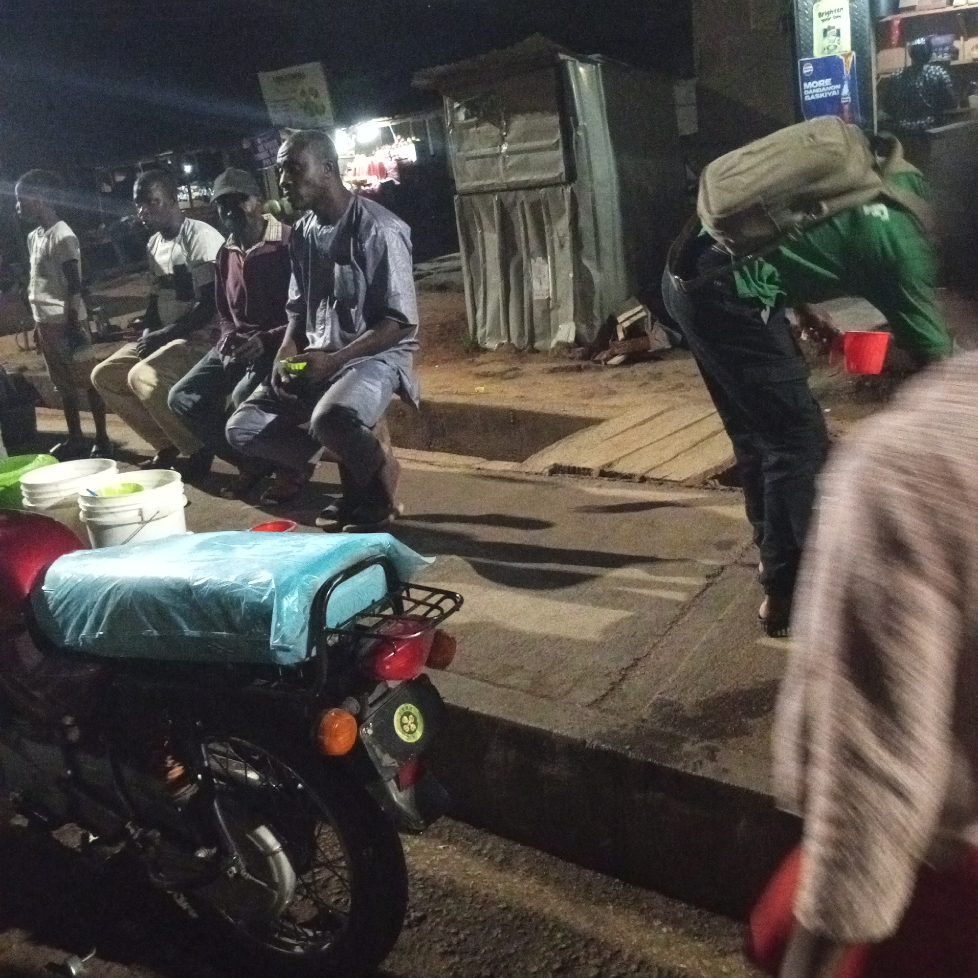 The image shows a street scene at night in Unknown, Unknown. Four men are sitting on a raised platform, likely a curb, on the left side of the frame. Two white buckets are placed next to them. A red motorcycle with a blue plastic cover over the seat is parked in the foreground. To the right, a man wearing a green jacket and a backpack is bent over, holding a red cup or container. A metal structure, possibly a stall or shop, stands behind the seated men. The lighting is dim, with illumination from overhead sources and nearby buildings. A sign is visible in the background above the metal structure. The foreground shows a section of pavement and a dark patch of ground. A person with a striped shirt is partially visible in the bottom right corner.