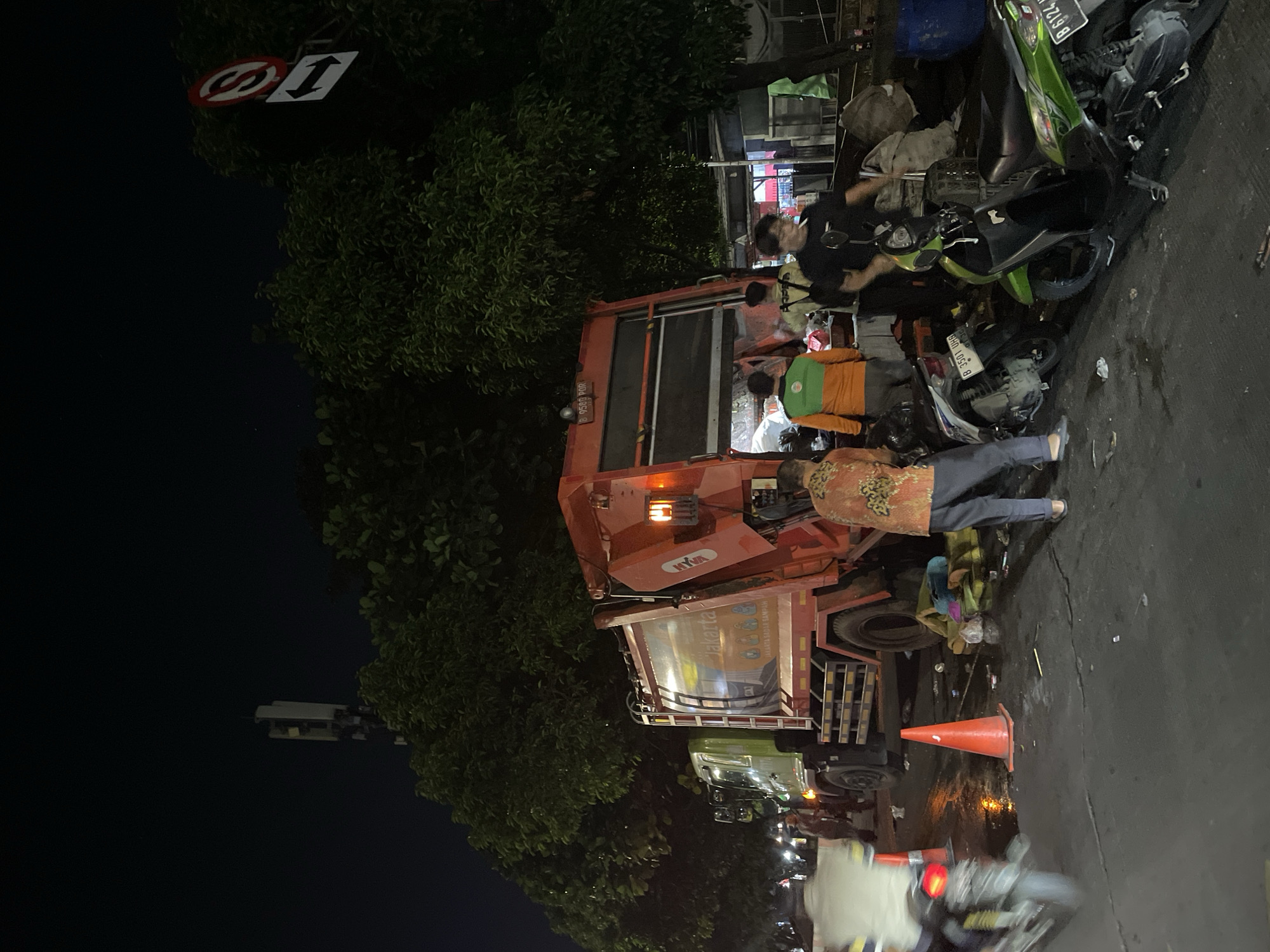 The image depicts a street scene in North Jakarta, Indonesia at night. A parked refuse collection truck, labeled "Jakarta", is illuminated by its front lights and surrounding streetlights. Several individuals are gathered around the truck and nearby motorcycles, which appear to be damaged and on the ground. A person in a green shirt is visible near the vehicles. An orange traffic cone is positioned on the road next to the truck. A "no entry" traffic sign is visible amongst some roadside trees. The scene suggests a traffic accident or incident involving the garbage truck and the motorcycles.