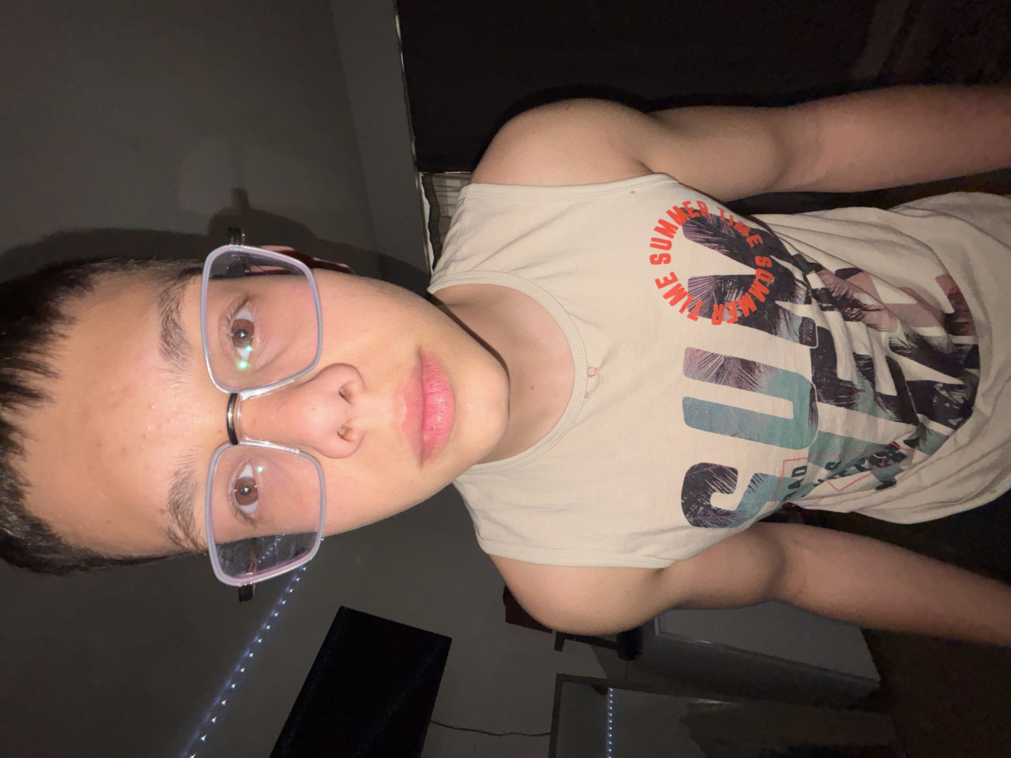 This rotated close-up portrait features a young individual, likely male, with short dark hair and light skin, wearing modern transparent-framed glasses. They have brown eyes and a neutral, direct gaze towards the camera. The individual's lips are slightly parted, revealing a light pink hue. They are dressed in a light beige sleeveless tank top, which features a prominent graphic design of palm trees in shades of blue, white, and black. The words "SUMMER TIME" are visible in orange lettering as part of the shirt's design. The background is dimly lit and largely dark, indicating an indoor setting, possibly a room at night or with low artificial lighting. Partial views of a dark object, potentially a screen or furniture, and a string of small, bright LED lights are discernible in the background. The individual appears to be posing for a selfie. This image was captured in Troyan, Bulgaria.