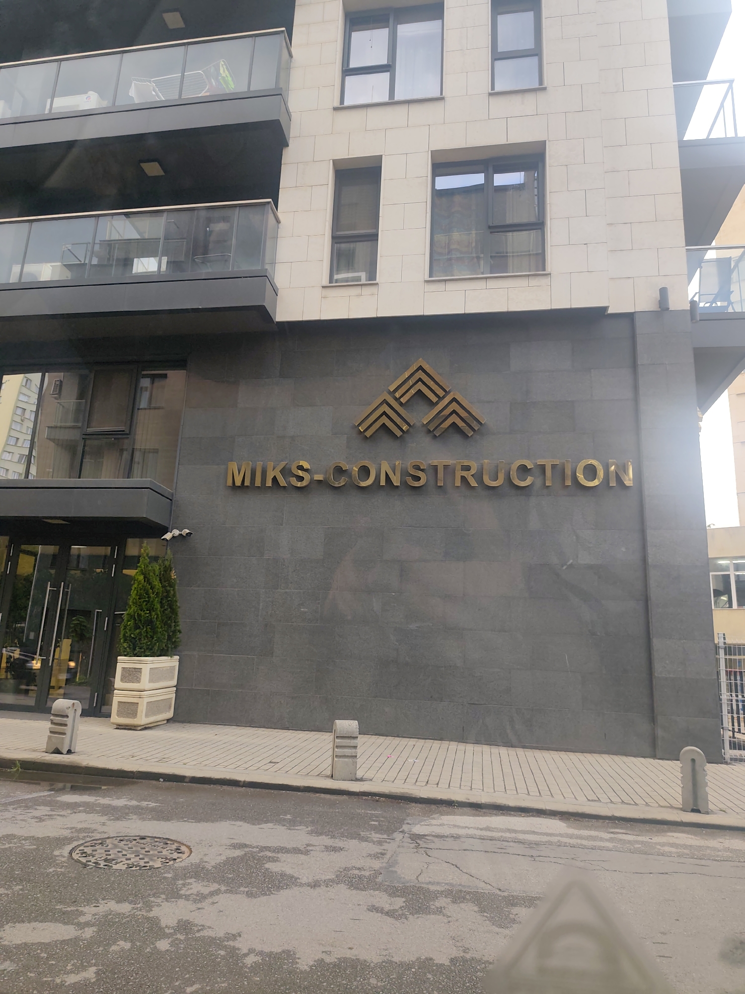 The image displays the street-level exterior of a modern building in Sofia, Bulgaria. The building's ground floor features a sleek, dark gray, textured stone facade, while the upper floors are constructed with lighter, possibly beige or off-white, block-tiled stone. Several windows with dark frames and balconies with glass railings are visible on the upper levels. One balcony prominently shows an air conditioning unit and a clothes drying rack, while another contains outdoor furniture like a table and chairs.
A large, golden sign is mounted on the dark gray facade. Above the text, there is a golden logo composed of three stacked, upward-pointing chevron shapes, reminiscent of rooftops or ascending arrows. The text clearly reads "MIKS-CONSTRUCTION" in golden, capital letters.
At the base of the building, a paved sidewalk, made of light-colored pavers, extends along the street. Concrete bollards with distinctive carved patterns are positioned along the edge of the sidewalk, separating it from the asphalt street. A circular manhole cover is visible on the street surface, which appears somewhat damp or marked. Directly in front of the building's main entrance, which features large glass doors, a well-maintained conical evergreen shrub is planted in a decorative, rectangular planter made of lighter stone.
The scene is devoid of people, suggesting a quiet moment. The lighting indicates it is daytime, with some glare reflecting off the golden sign. The overall impression is one of a contemporary urban environment.