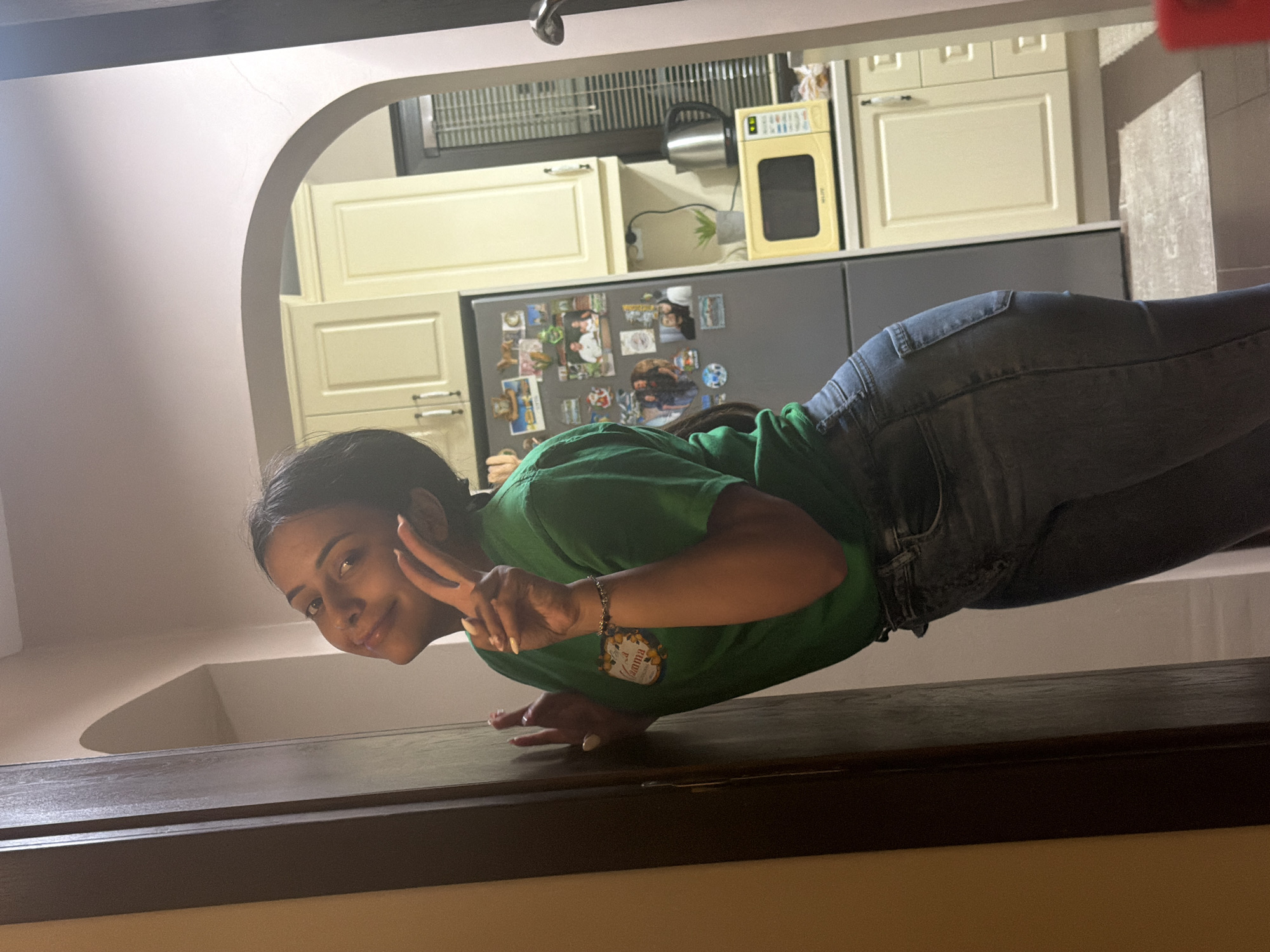 An indoor photograph captures a young woman in a dynamic balancing pose, possibly a planche or frog stand, on a dark wooden surface in the foreground. She is positioned horizontally relative to the viewer, wearing a green t-shirt and blue jeans. The woman looks directly at the camera with a cheerful smile, forming a peace sign with her left hand. Her dark hair is styled back, and she wears a beaded bracelet on her left wrist. A round sticker on her t-shirt features the text "La Mamma" alongside an illustration of lemons.
Behind the woman, through an arched doorway, a well-lit kitchen is visible. The kitchen features light cream-colored upper and lower cabinets with silver bar handles, a grey refrigerator extensively decorated with various photos and magnets, and a dark countertop. On the counter, a stainless steel electric kettle, a small potted plant, and a yellow compact appliance, possibly a 'Morphy Richards' toaster oven or air fryer, are arranged. The overhead area above the cabinets shows a metal grate, possibly for ventilation. The image is shot from a low angle, emphasizing the woman's athletic pose against the backdrop of a modern home kitchen. This lively scene takes place in Shumen, Bulgaria.