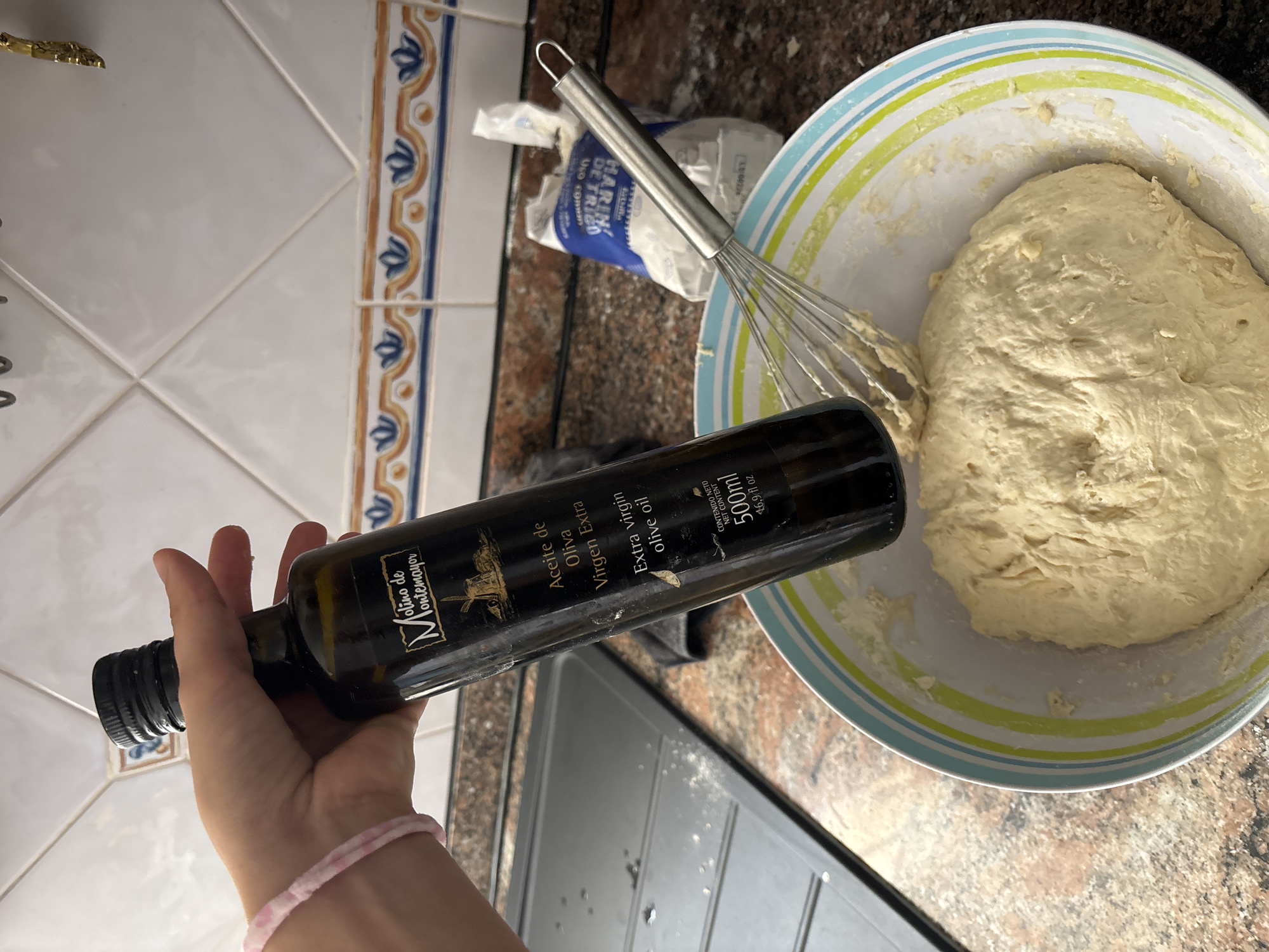 The image shows a hand holding a dark glass bottle of extra virgin olive oil. The bottle has a label with "Molino de Montemayor" and text in Spanish and English indicating it is "Aceite de Oliva Virgen Extra" and "Extra virgin olive oil," with a net content of 500ml. The hand is wearing a light pink bracelet. In the background, there is a kitchen counter with a stainless steel sink, a bag of flour, and a metal whisk in a bowl with dough. The kitchen has white tiled walls with a decorative border. The scene depicts food preparation. There is no indication that this scene is located in the city of Orihuela, Spain.